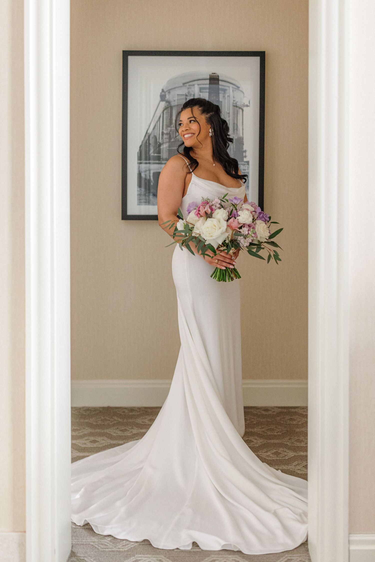 Elegant bride in fitted white wedding gown holds romantic bouquet while posing in doorframe.