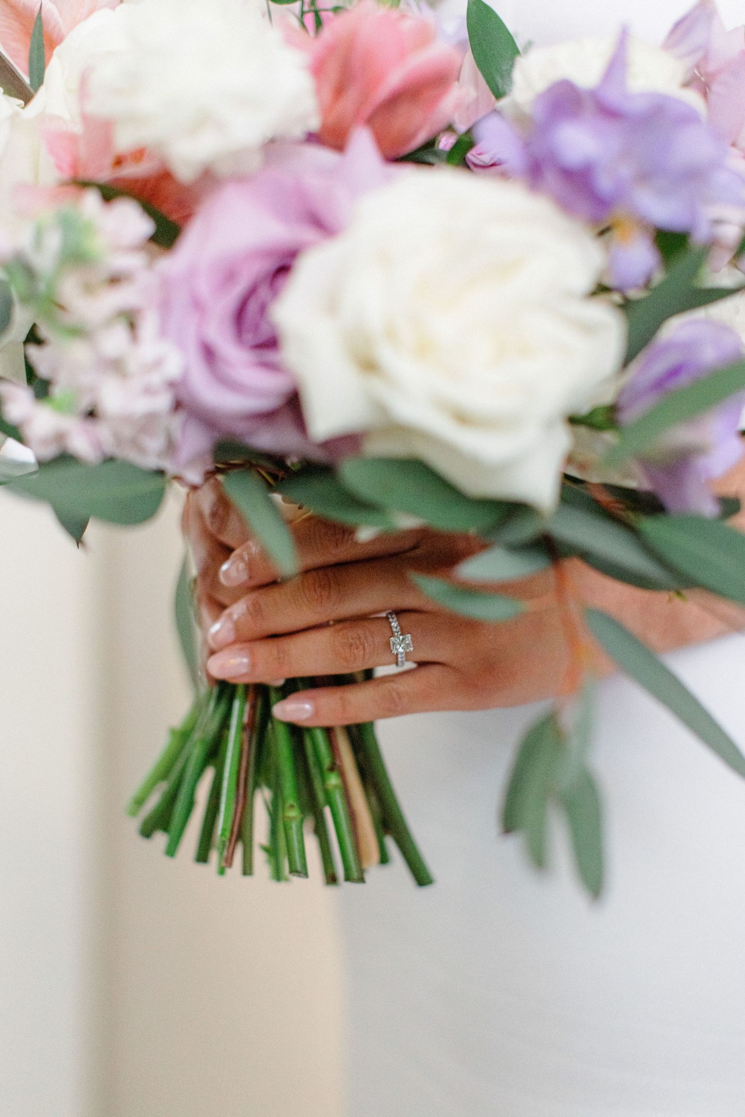 A delicate bridal bouquet of pink, purple, and white flowers held by hands showcasing an engagement ring.