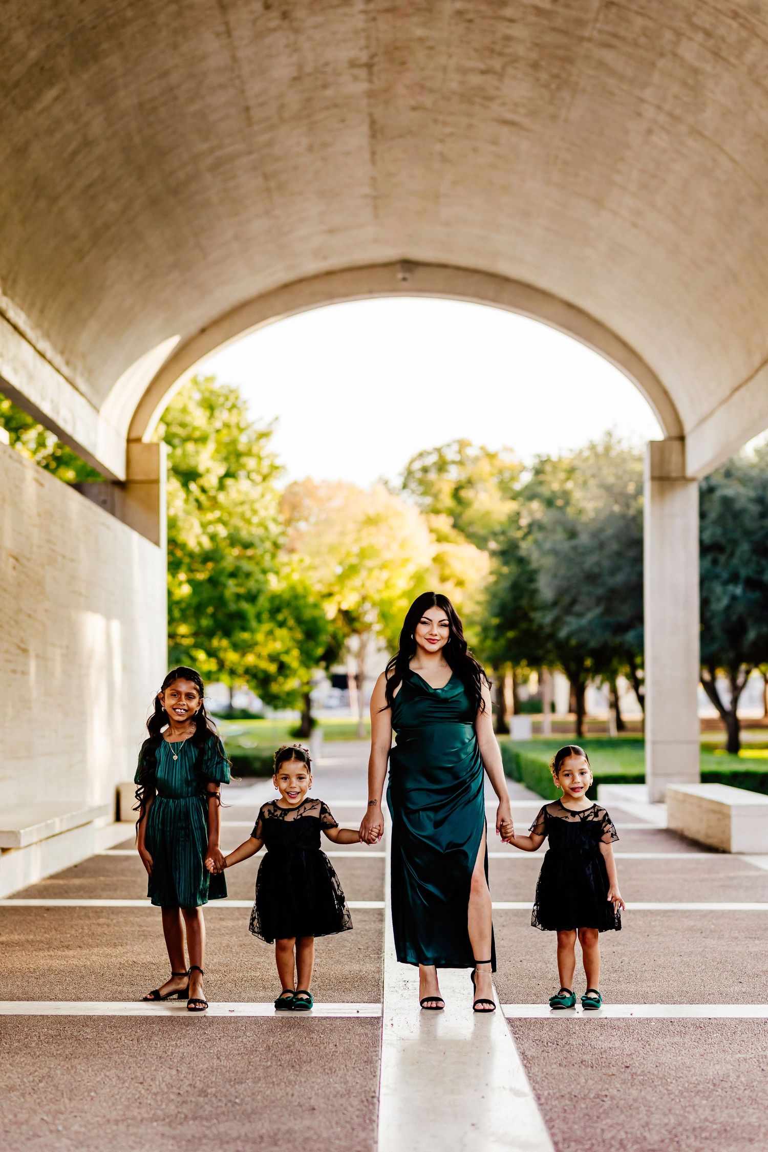 Four people in elegant dresses stand symmetrically under a concrete archway in a park.