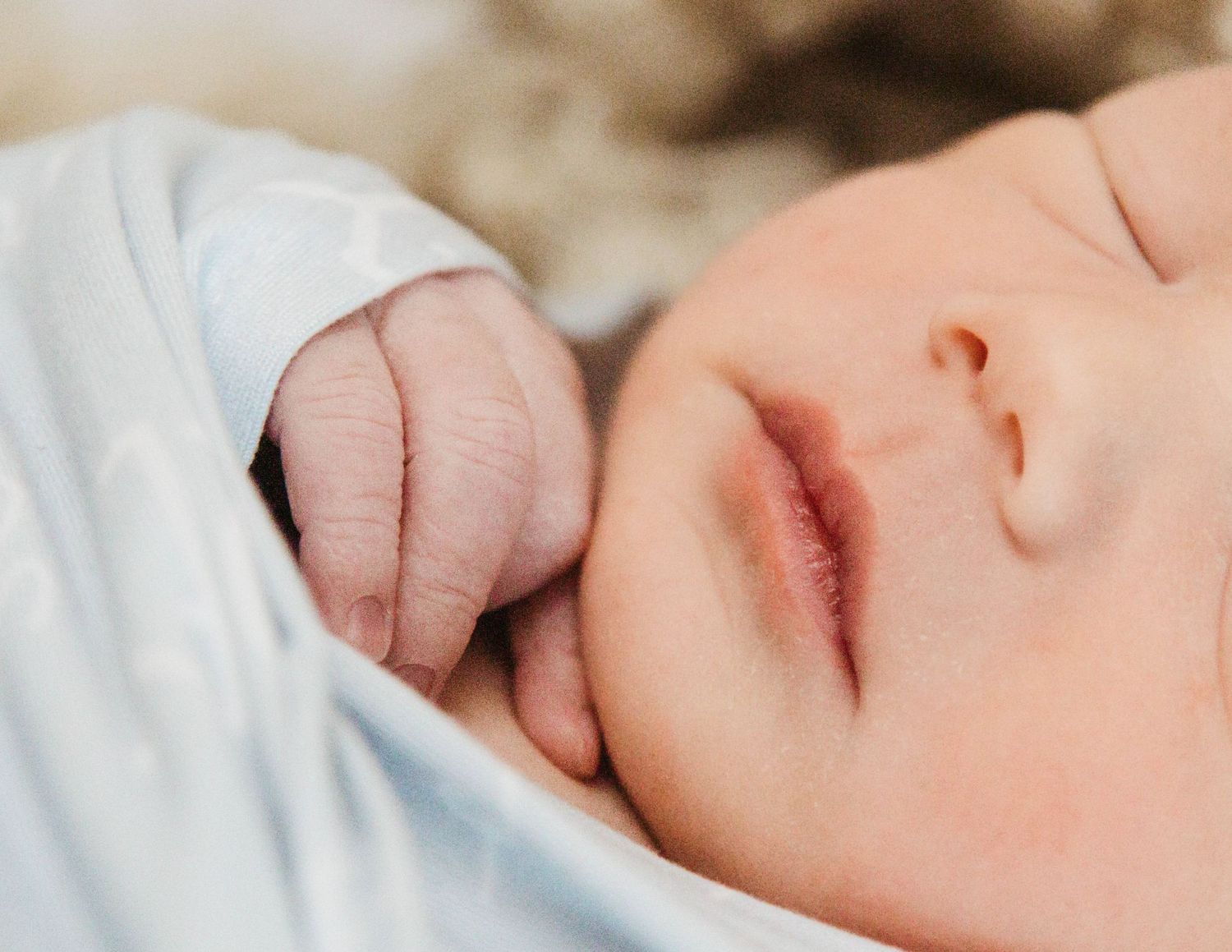 A sleeping newborn baby wearing a white outfit rests peacefully with their small hand curled up.