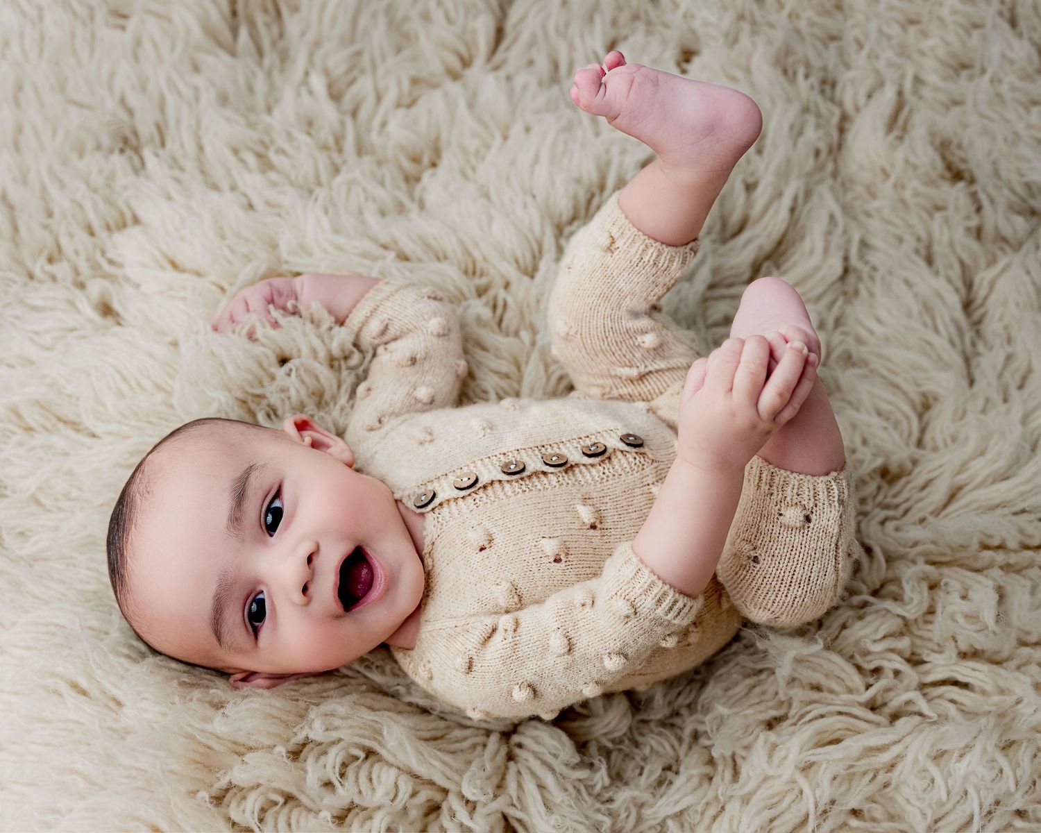 Baby in cream knit outfit lying on fluffy white rug with tiny feet up.