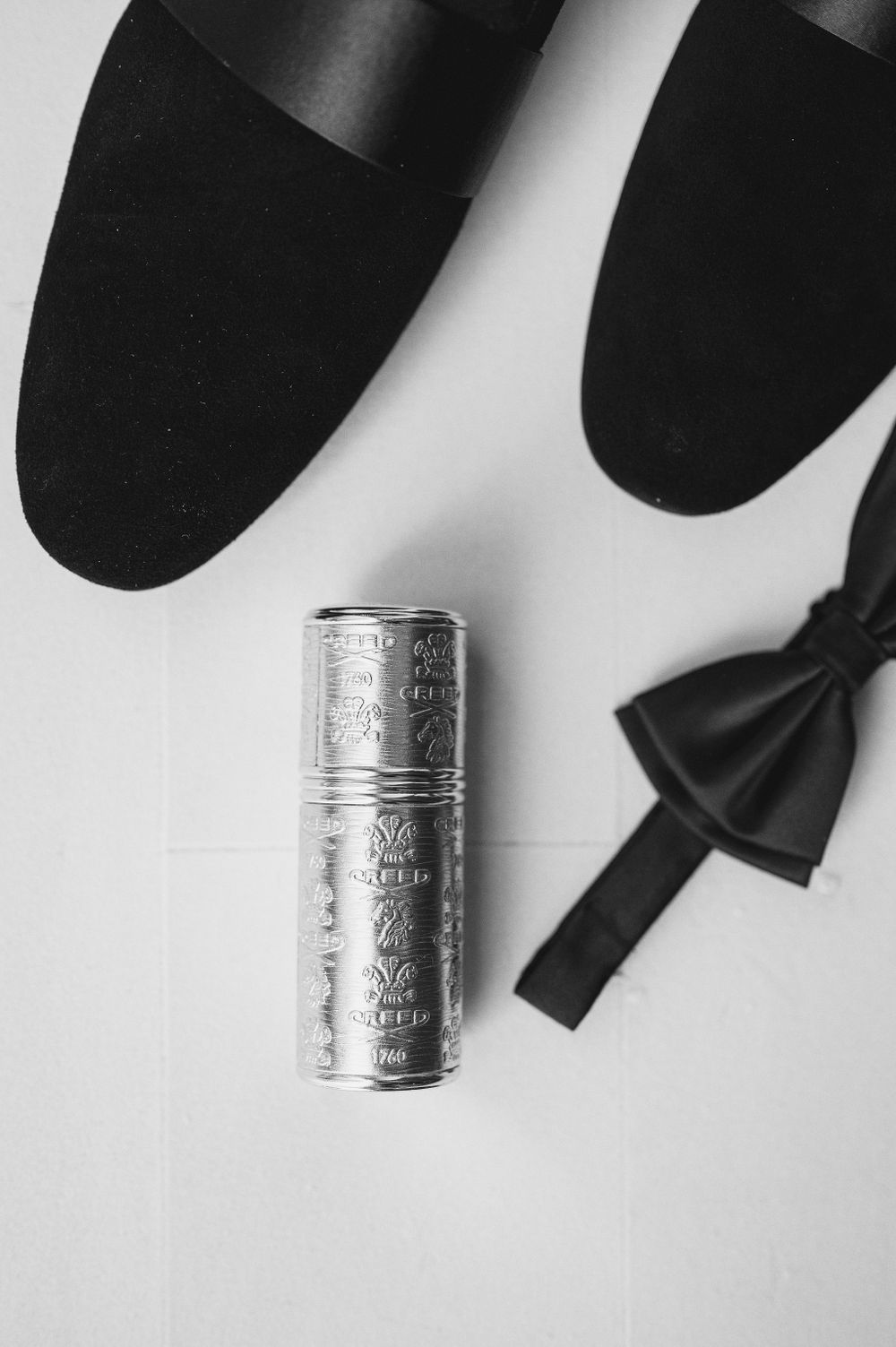 Black dress shoes, bow tie, and silver metal can arranged on white surface.