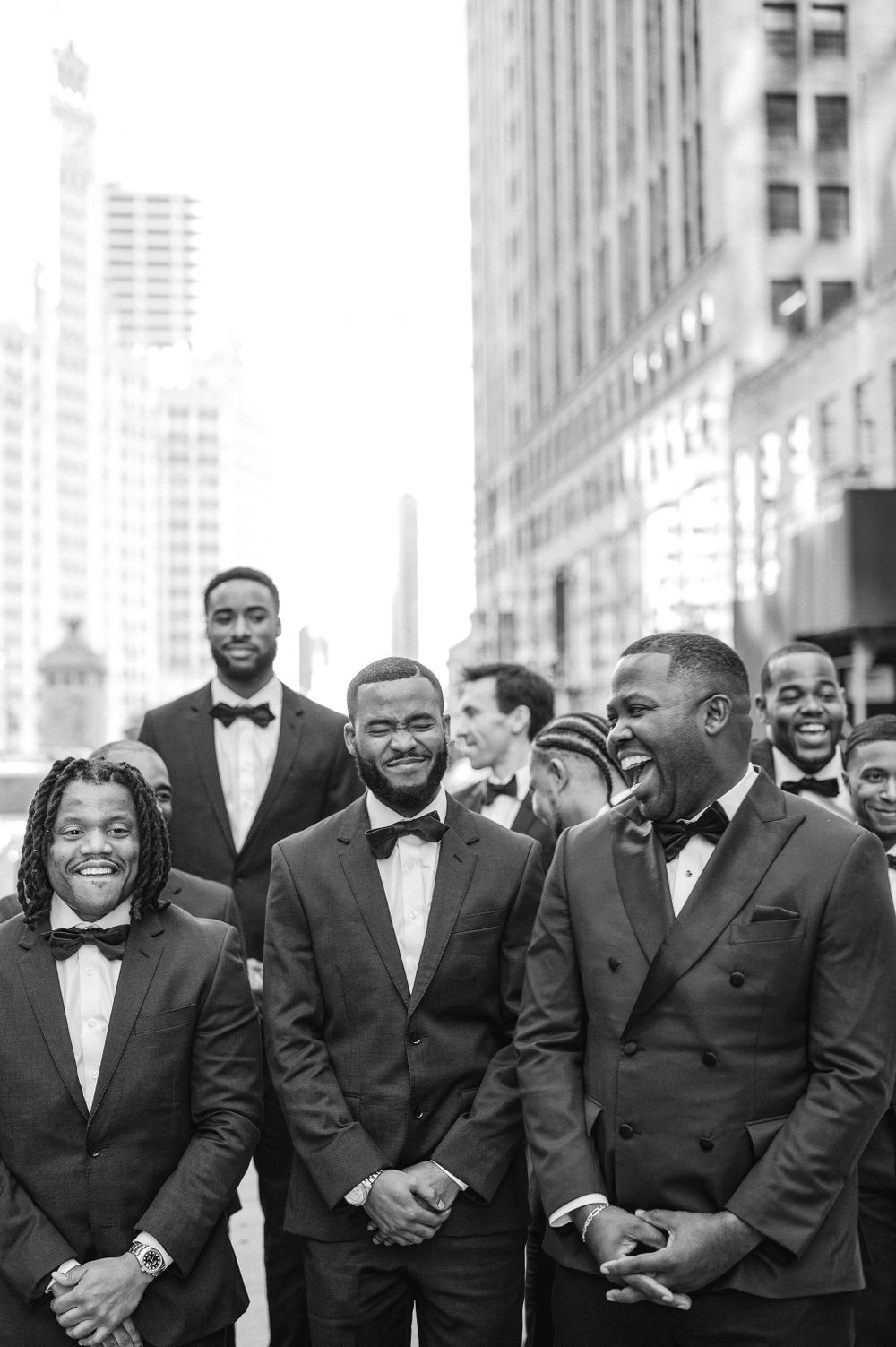 Groomsmen in black tuxedos share a candid moment in front of city buildings.