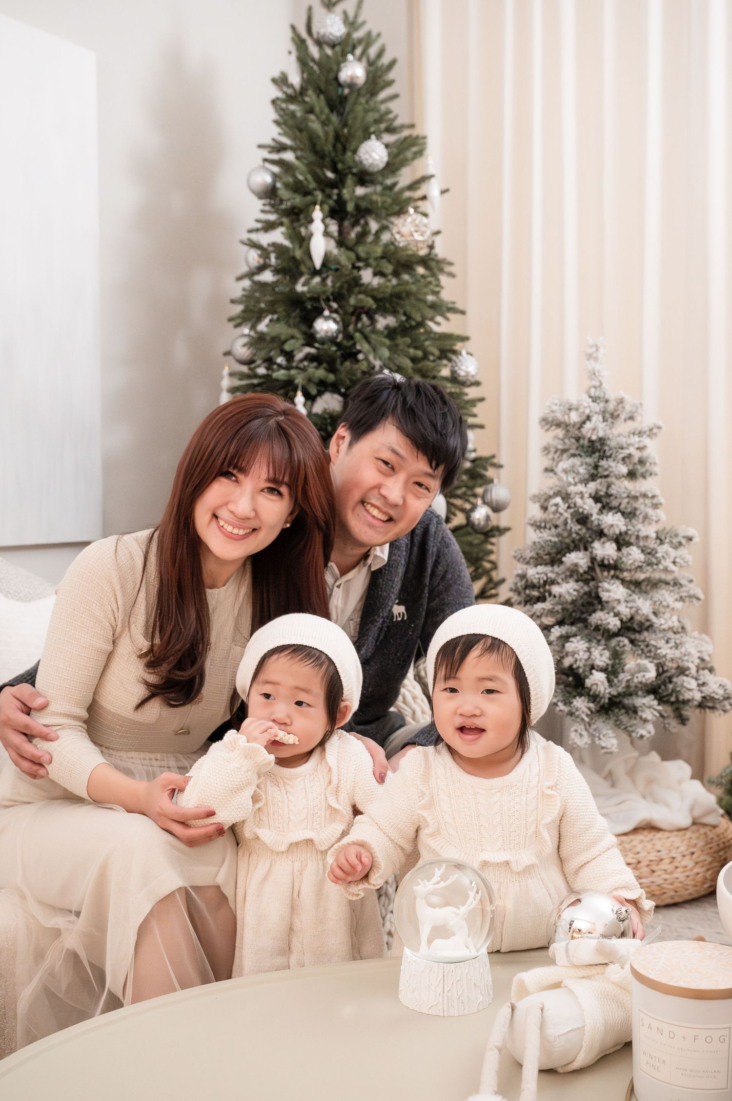 Family lounging together by decorated Christmas trees with twins in matching white outfits and winter decor.