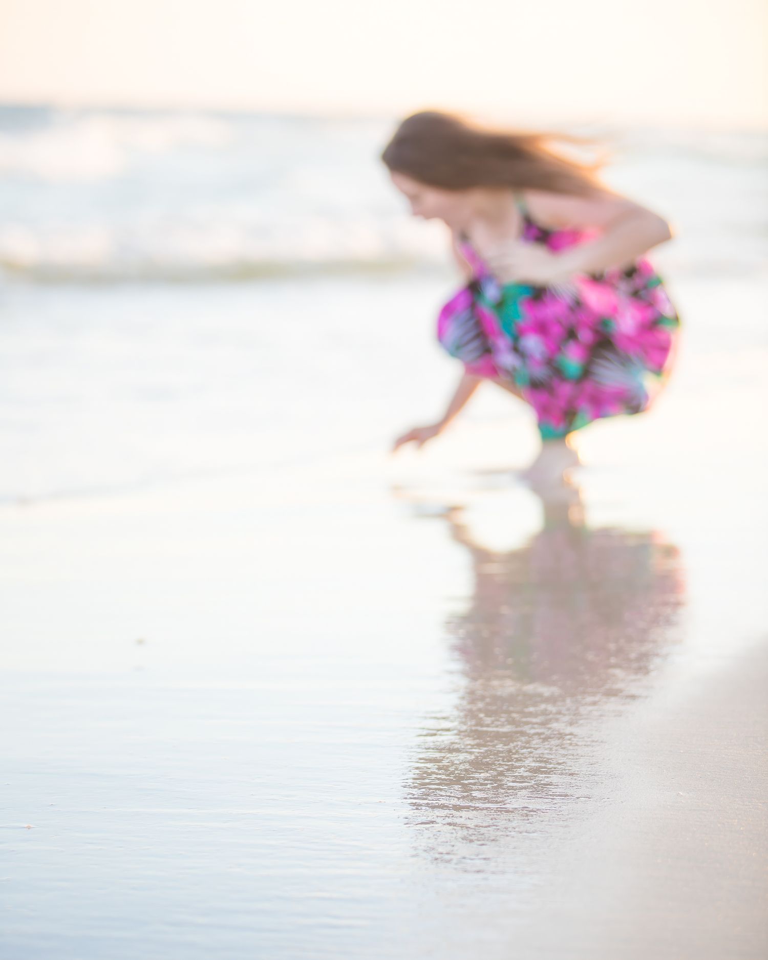 Dreamy soft focus image of someone in a floral dress bending down at the beach shore during sunset.