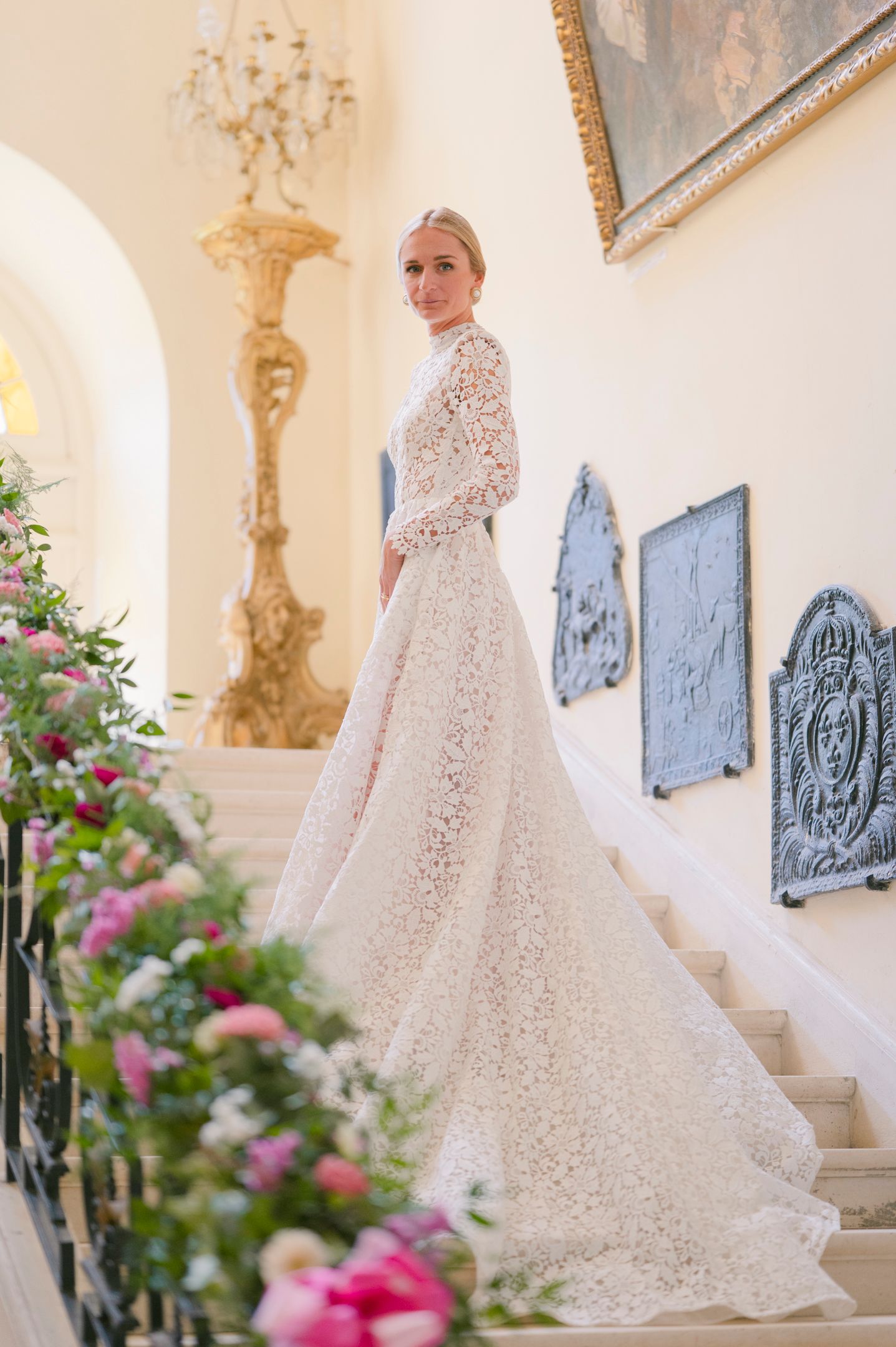 An elegant bride poses on ornate stairs with lace gown and trailing train amidst pink flowers.