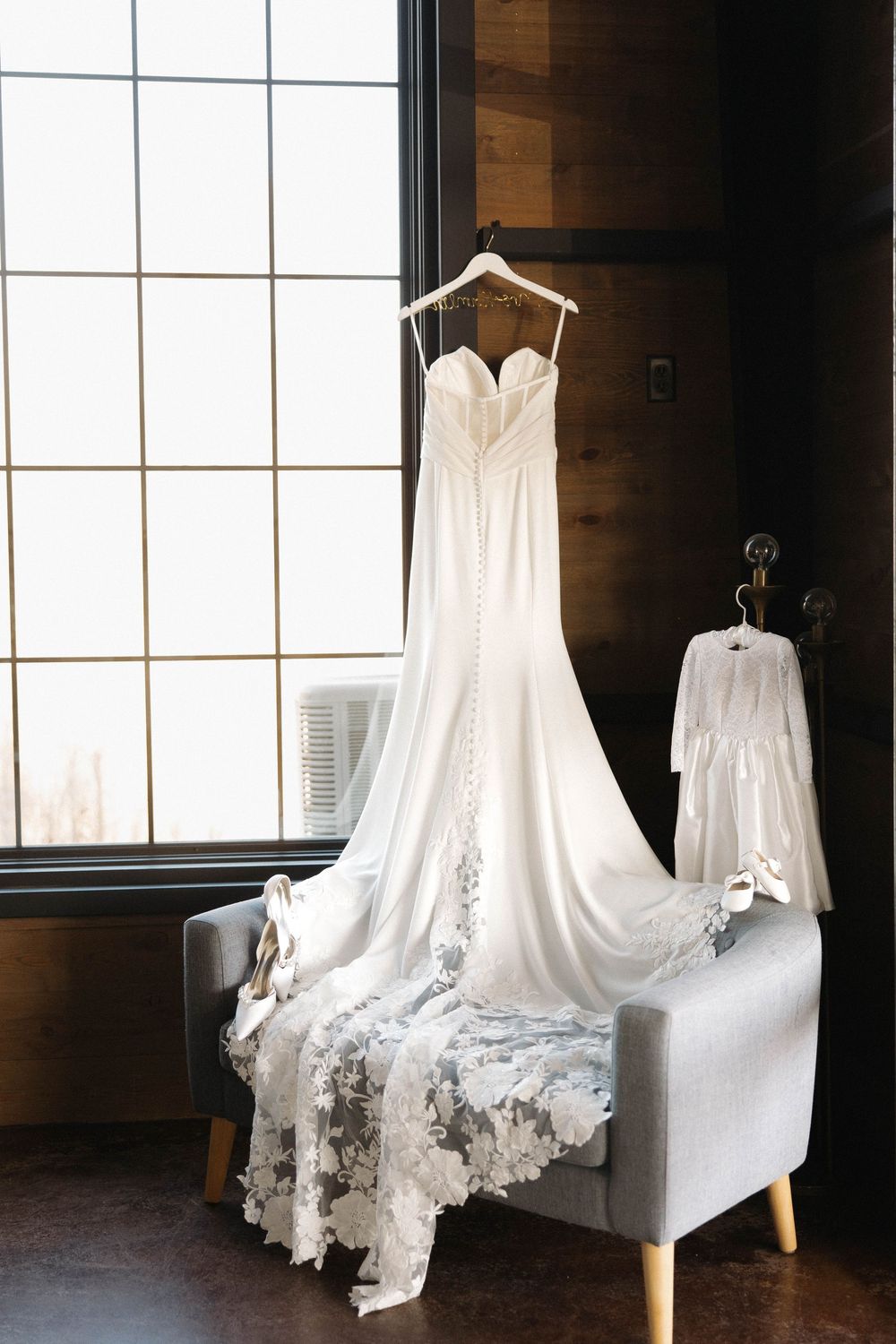 A white lace wedding dress with sweetheart neckline hangs elegantly on a hanger near a bright window.