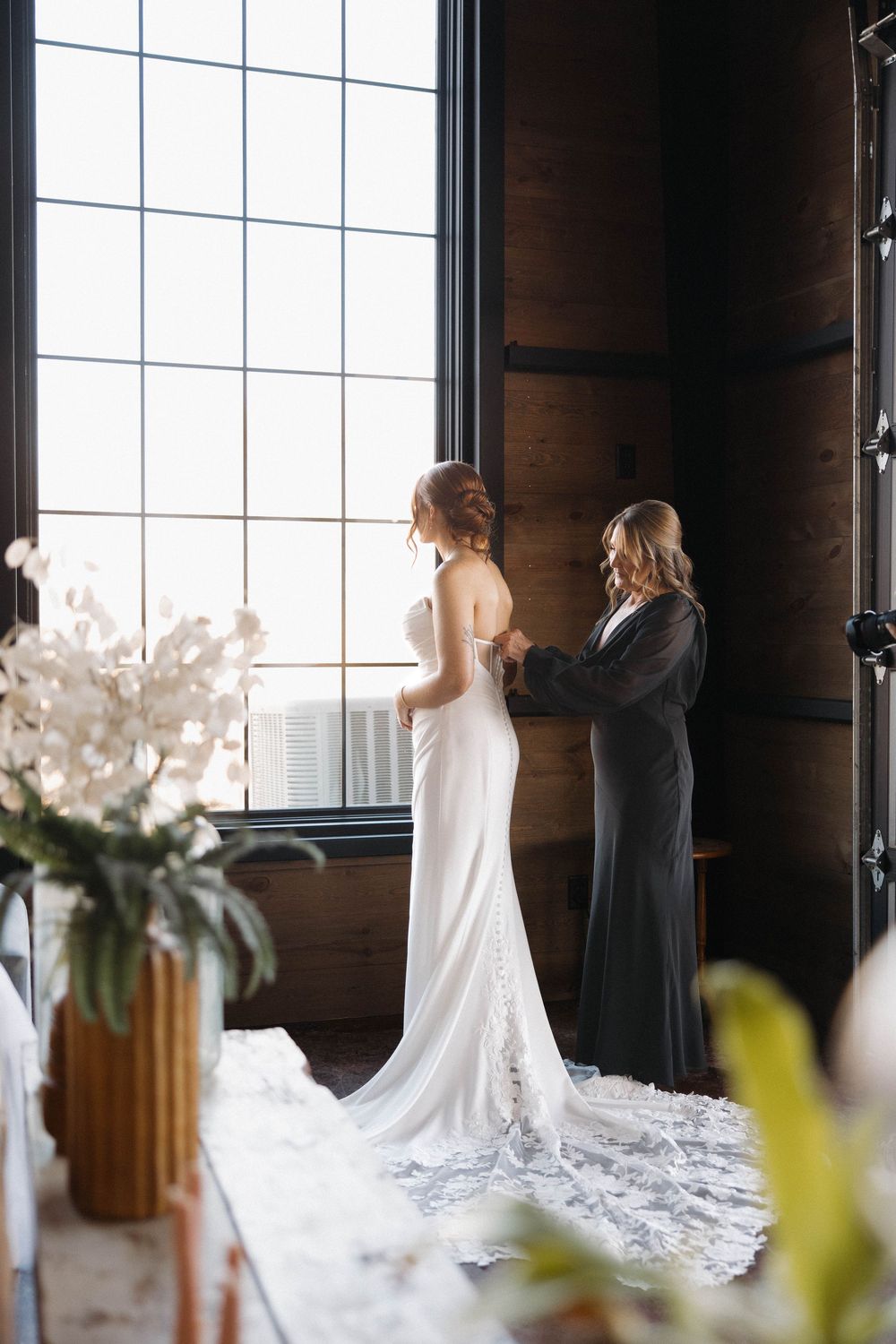 A bride in a flowing white gown with a long train stands by large windows in an elegant indoor setting.