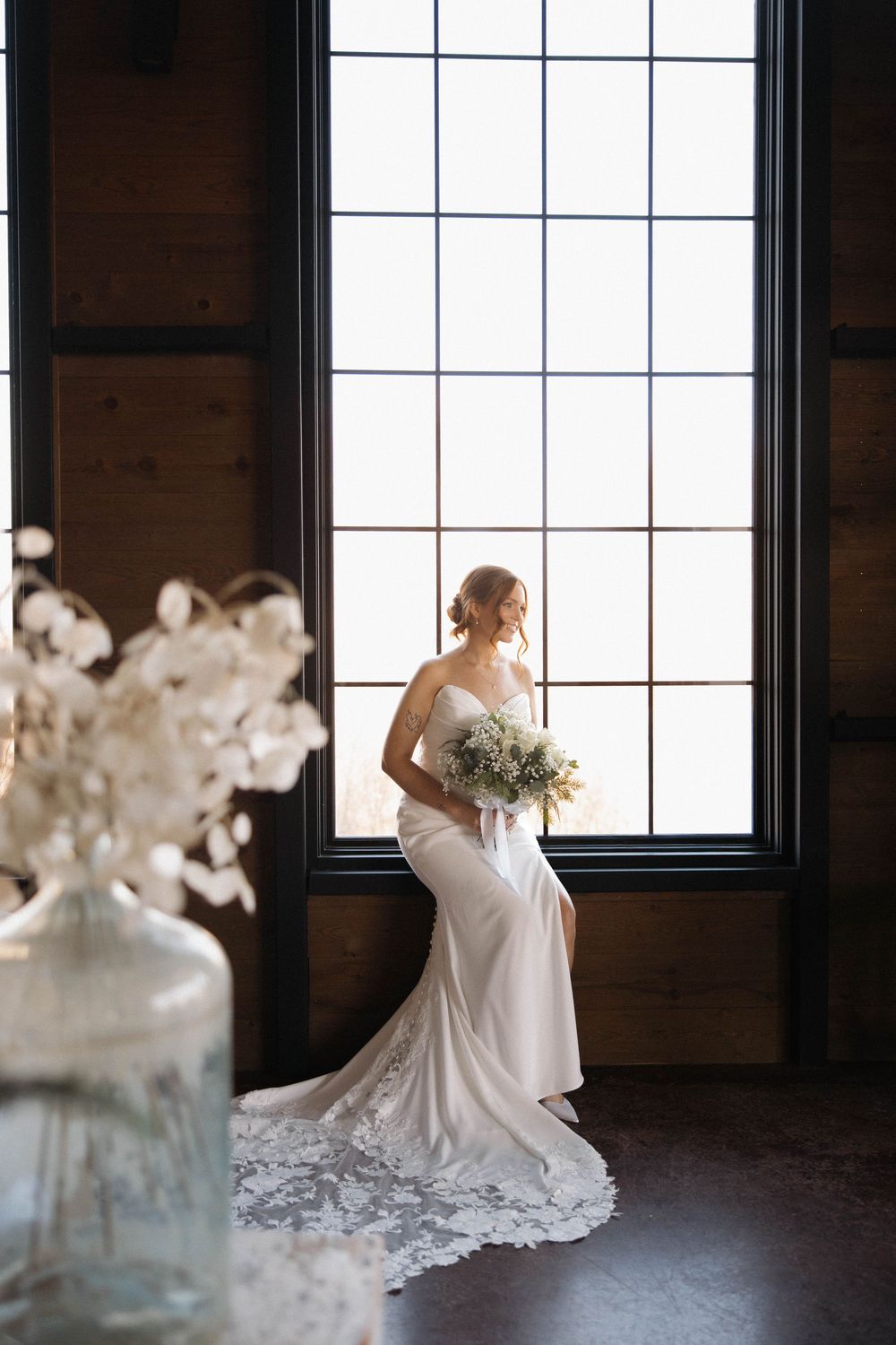 Elegant bride in fitted lace gown poses by large window with bouquet in rustic indoor setting.
