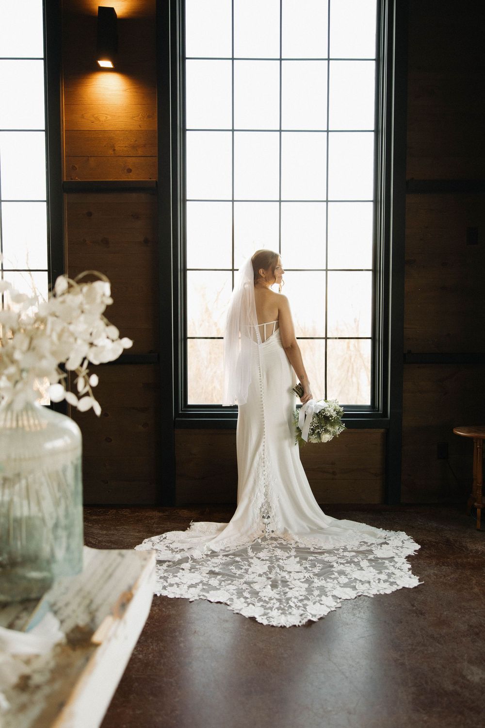 A bride in a white lace wedding gown with a dramatic cathedral train stands silhouetted against bright windows.