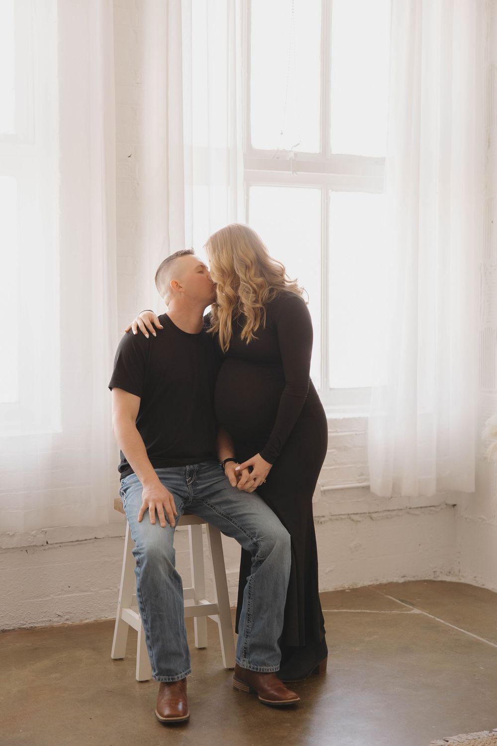 Intimate indoor photoshoot with two people in black clothing sitting on white chairs against bright windows.