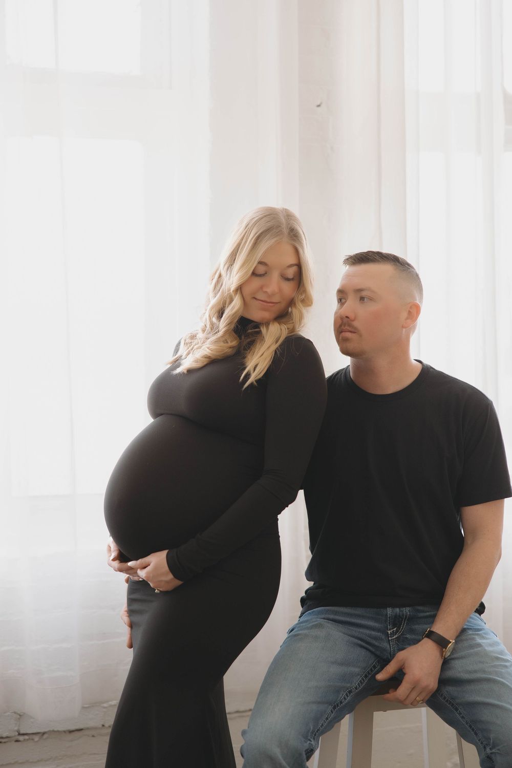 Couple in matching black outfits posing together in bright white studio setting.