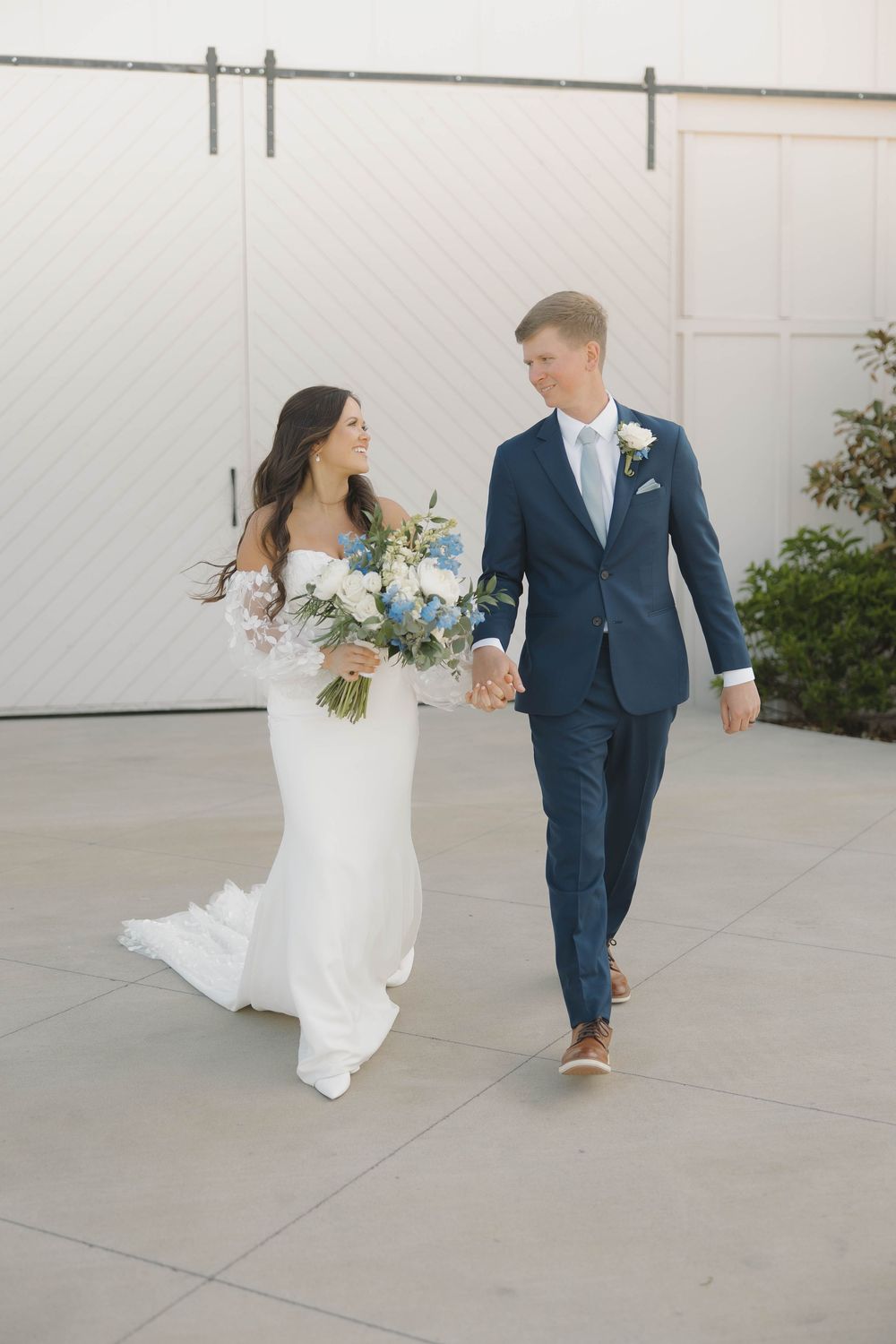 A bride in a fitted white gown and groom in navy suit walking together and holding hands at an outdoor venue.