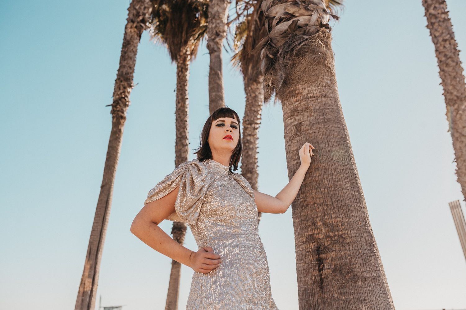 A woman wearing a glittery dress looks away while leaning on a palm tree in Santa Monica.