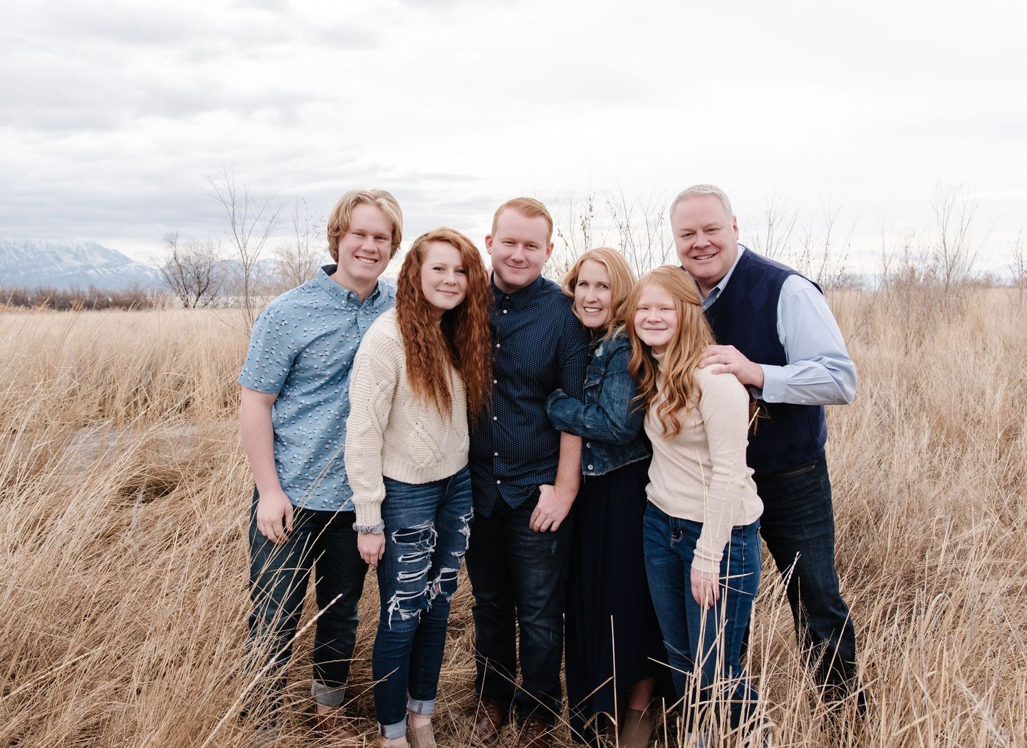 Family group poses together outdoors in tall grass with mountain landscape behind them.