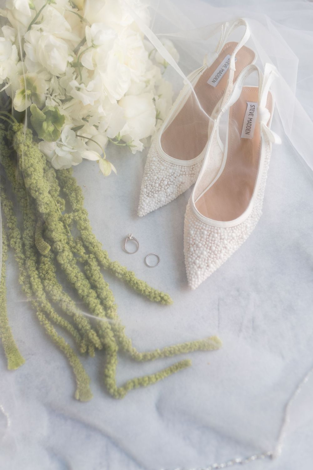Elegant sparkly white bridal slingback heels with wedding rings and white floral bouquet on marble background.