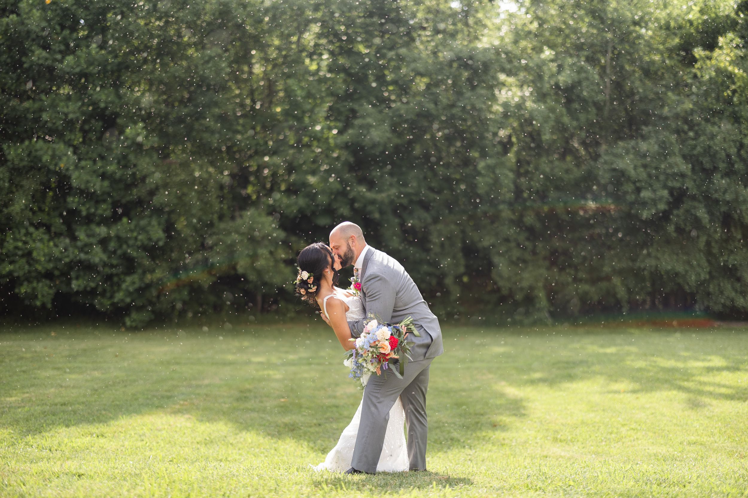 A couple embraces on a sunny lawn in front of trees during a romantic outdoor wedding photo session.