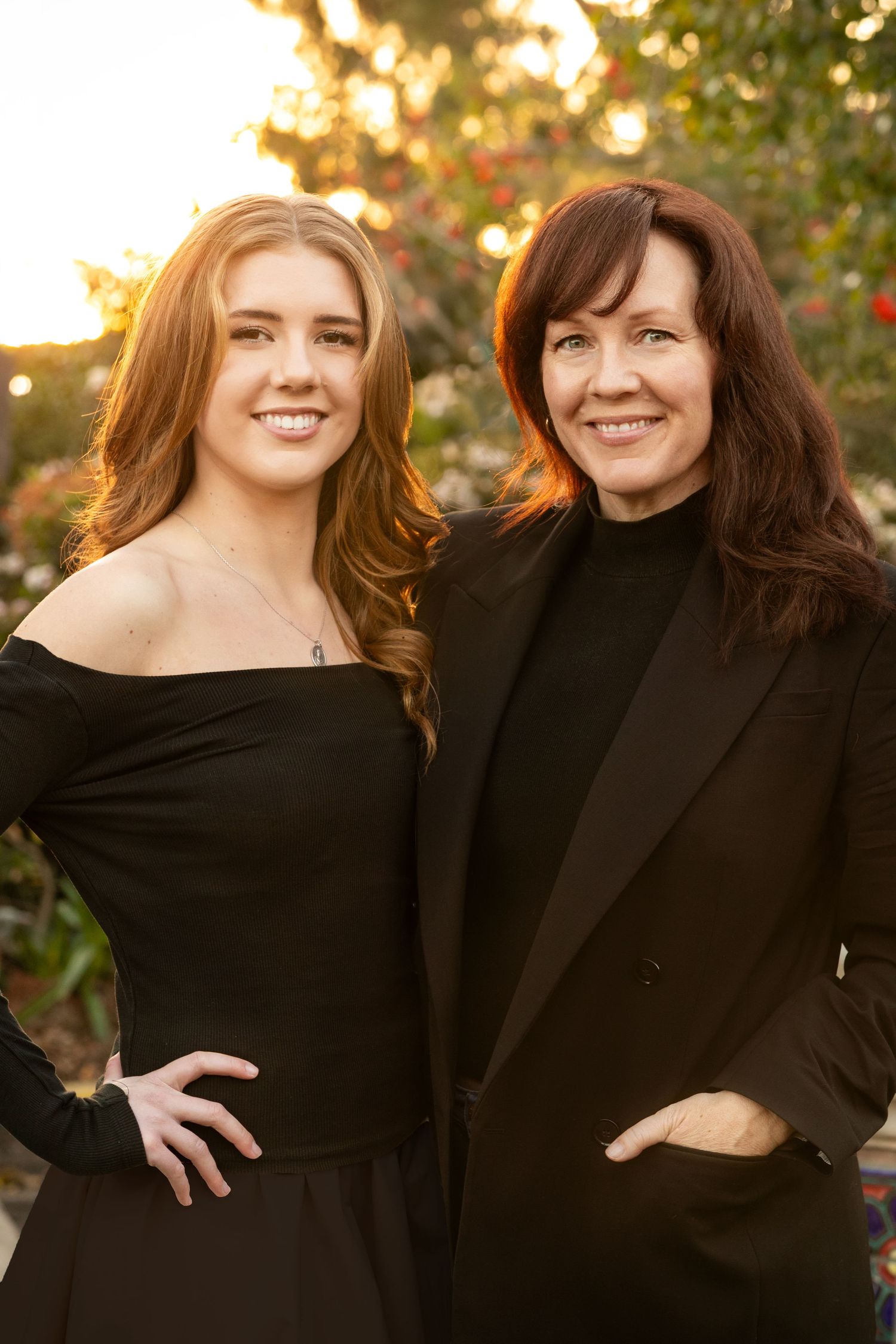Two people in matching black outfits smile warmly while posing together outdoors on a sunny day with bokeh background.