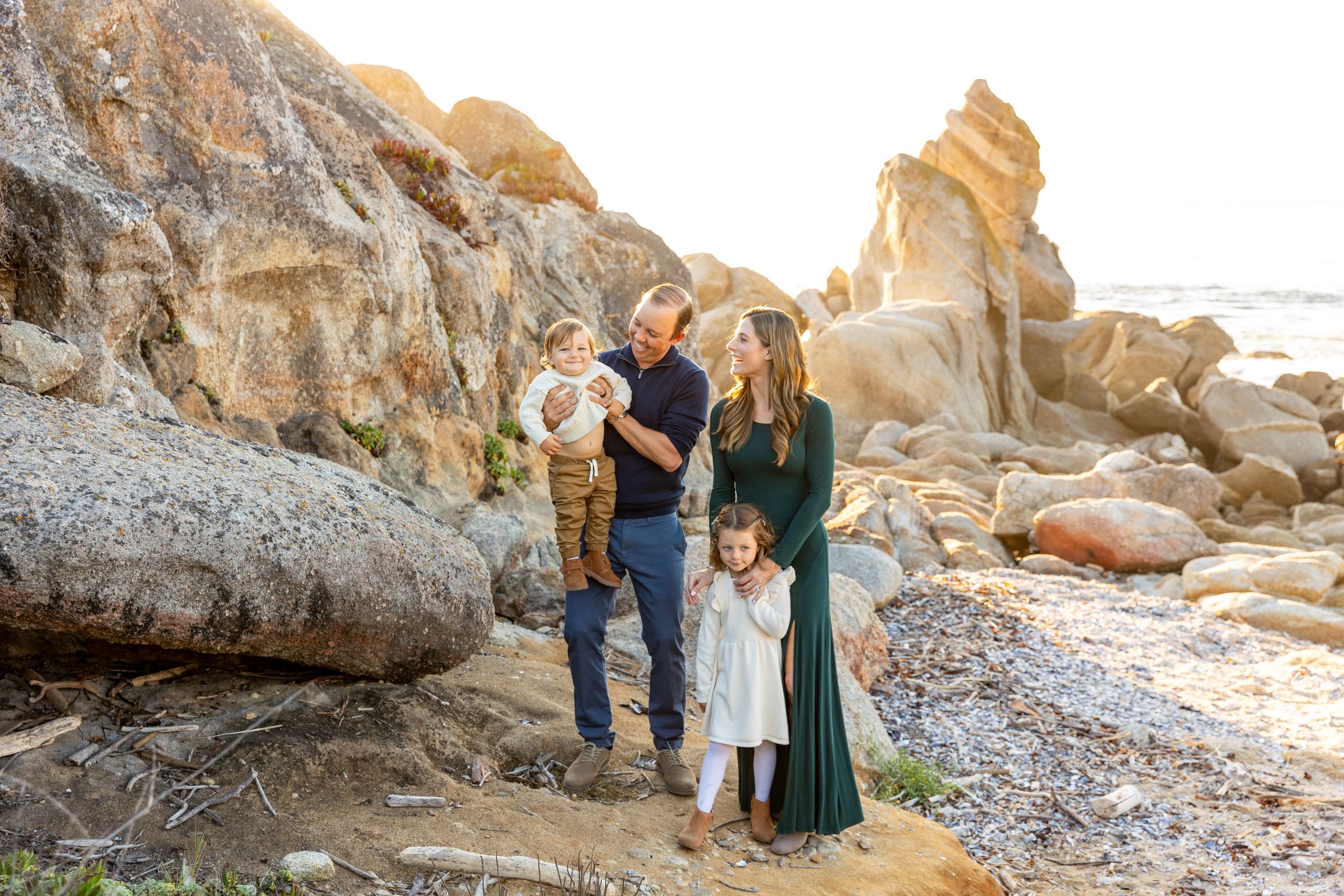 A family stands together on Carmel Beach at sunset with rocks in the background with Rachel Zee Photography.