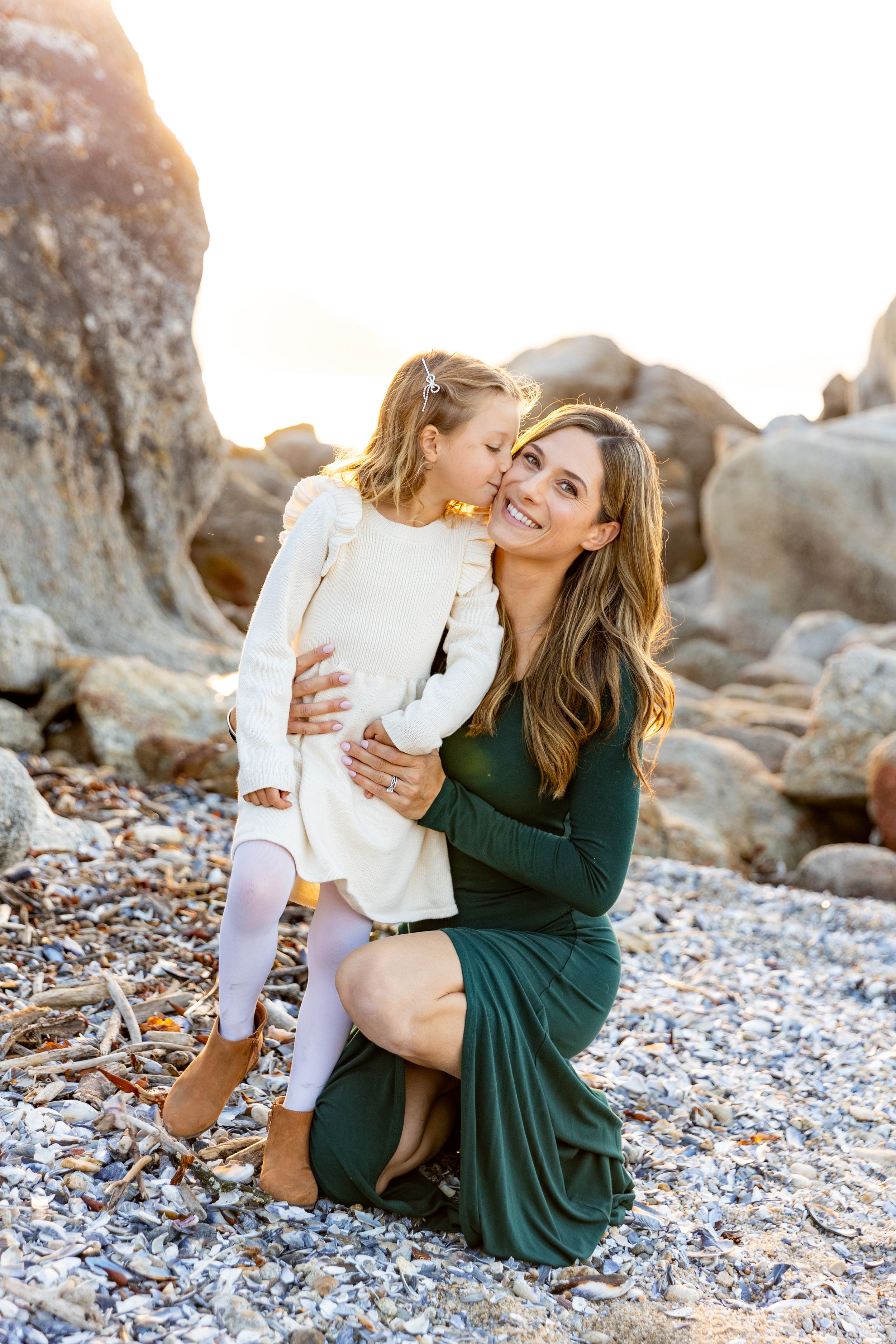 Mother and daughter share a tender moment together on a rocky beach during golden hour at sunset for family pictures.