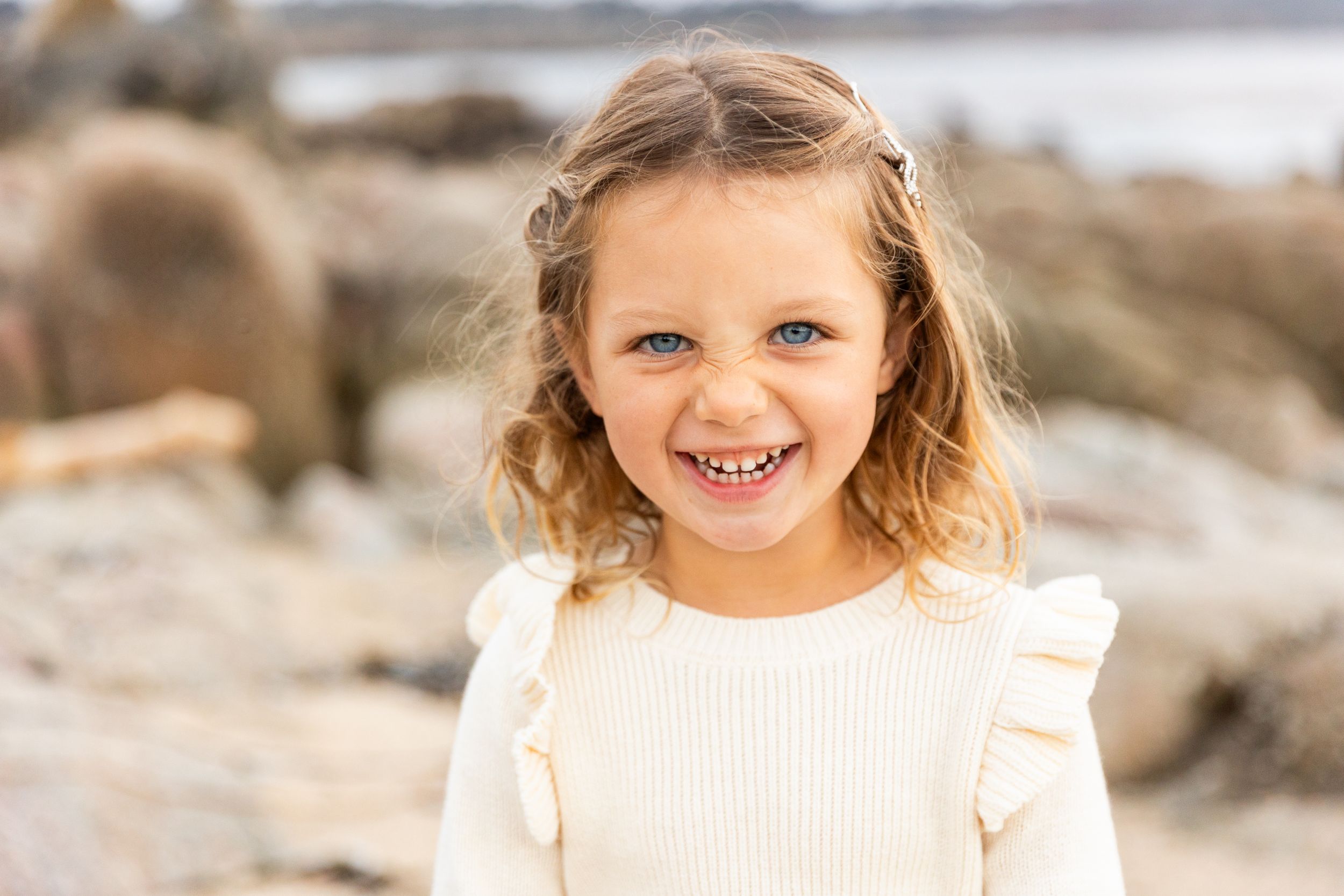 A child with wavy hair and a bright smile poses for a portrait at the beach.