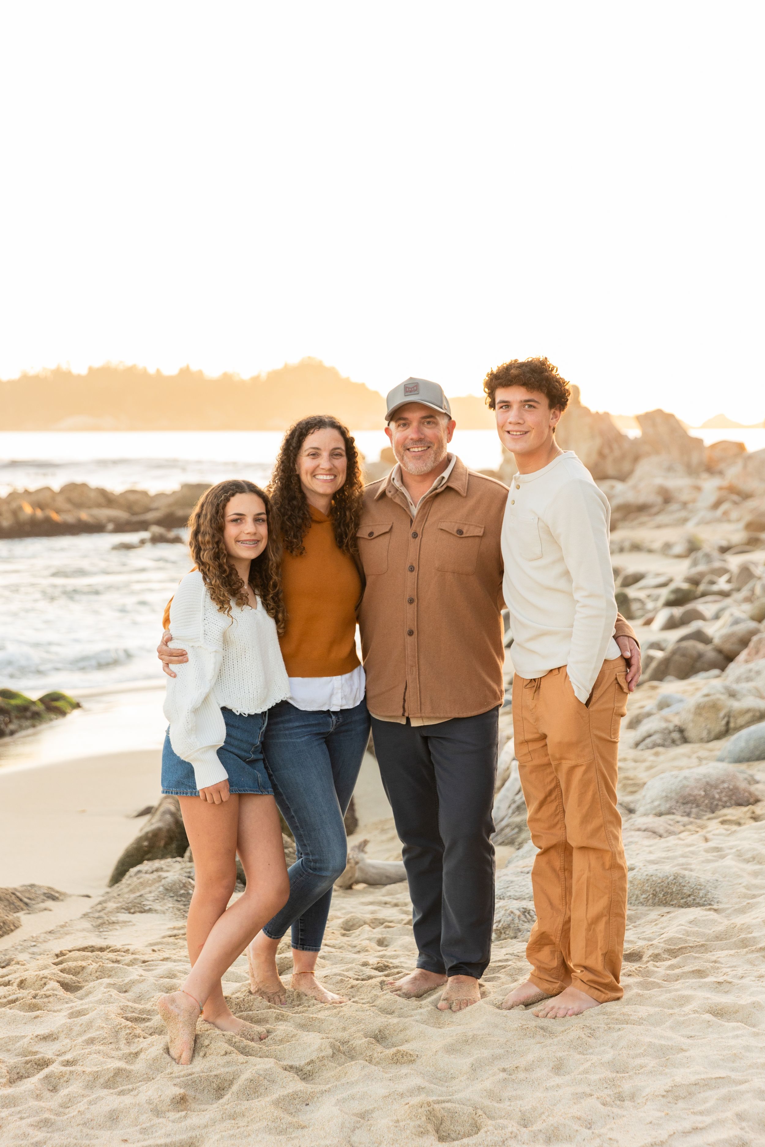 A family poses together on a sandy beach at sunset with rocky California coastline in the background.