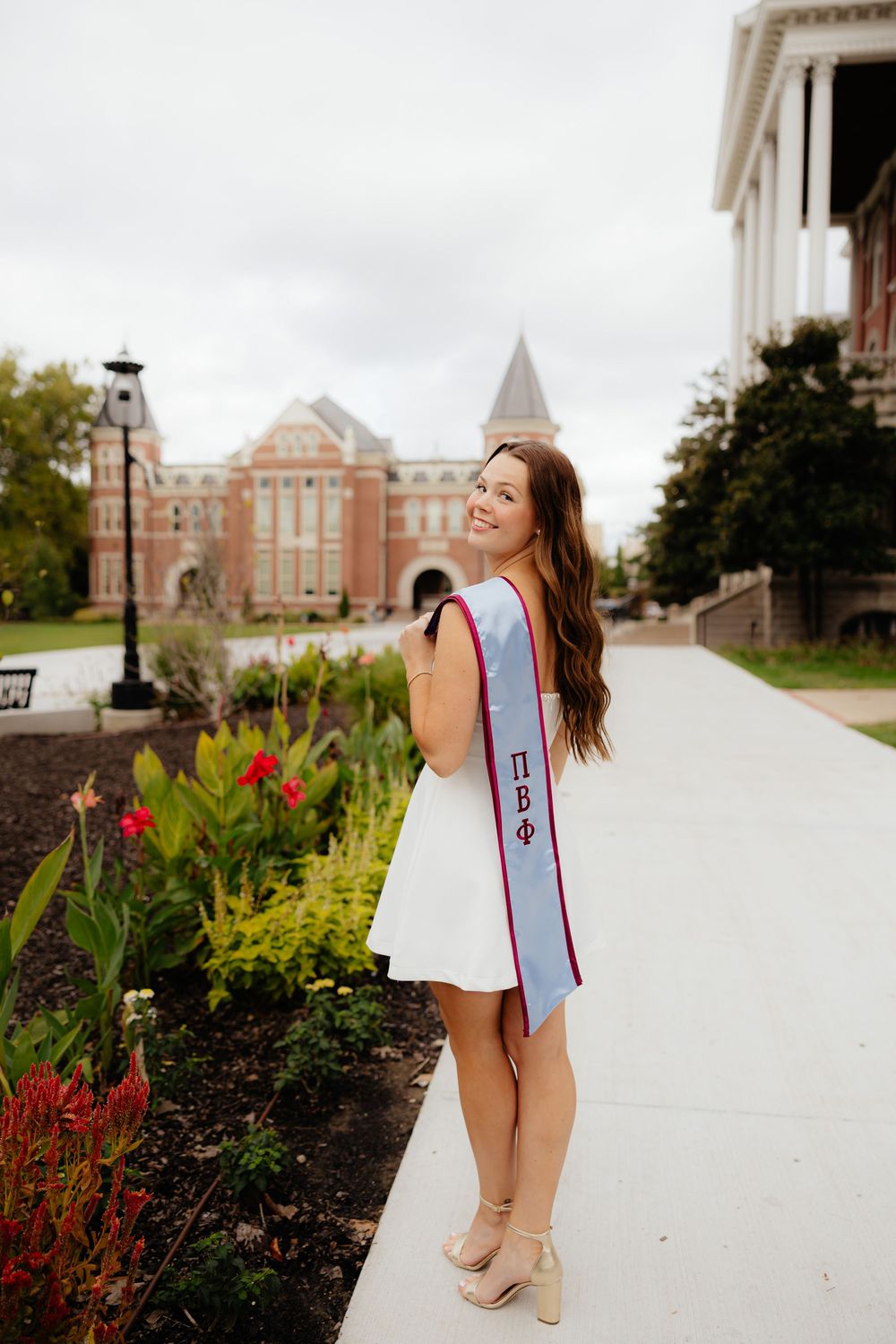Graduate poses in white dress with graduation sash on campus with red brick buildings and flowers in background.