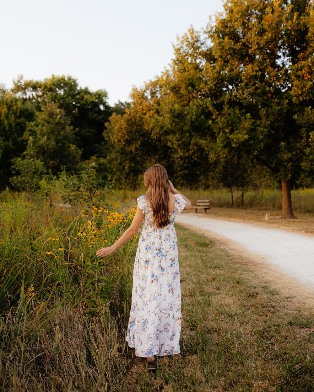 Flowing dress catches evening light on nature trail with autumn trees.