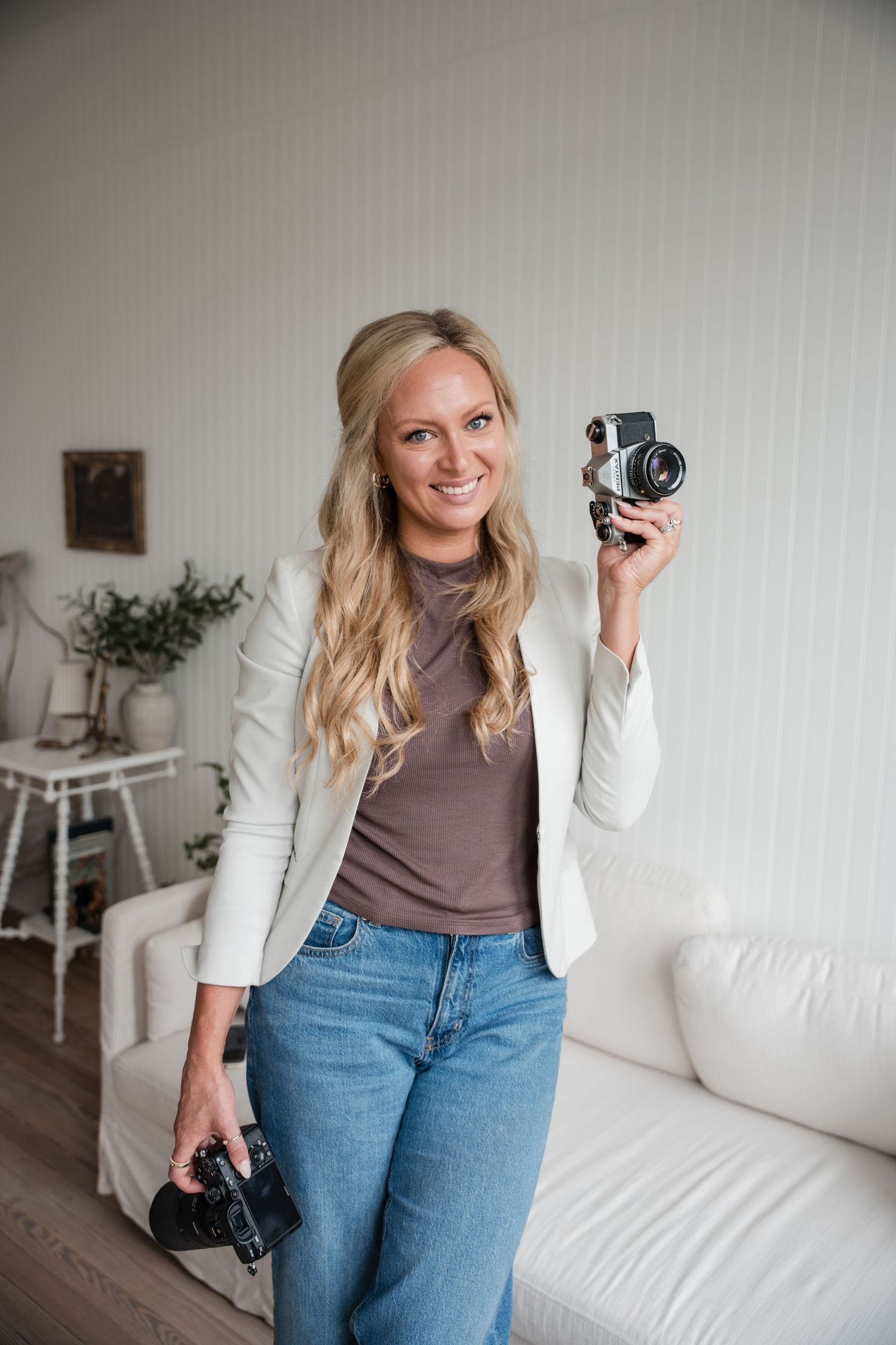 A photographer in a purple tank top poses with a professional camera against a grey background.