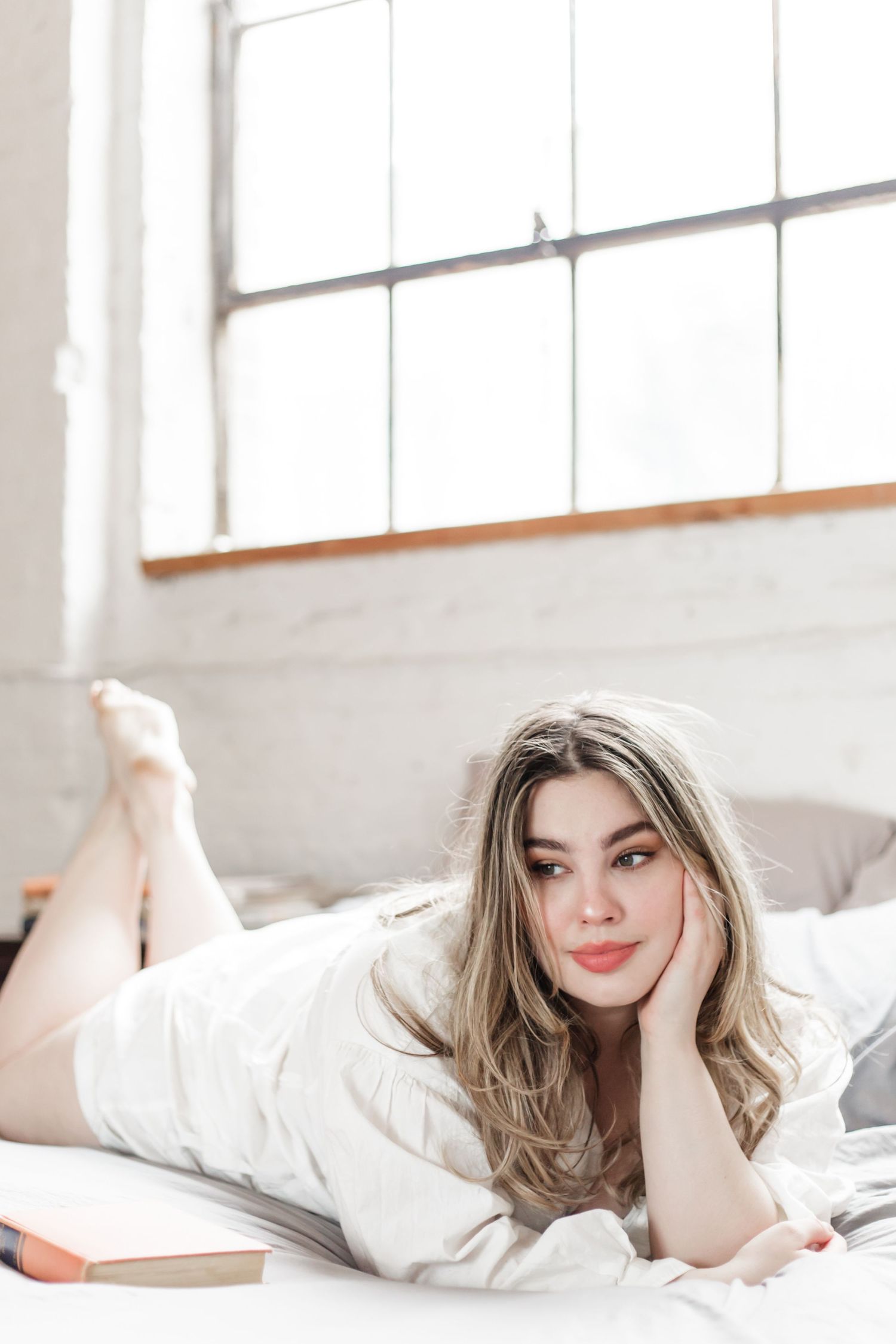 Soft boudoir shot of woman in white lounging on bed in studio with natural window light.