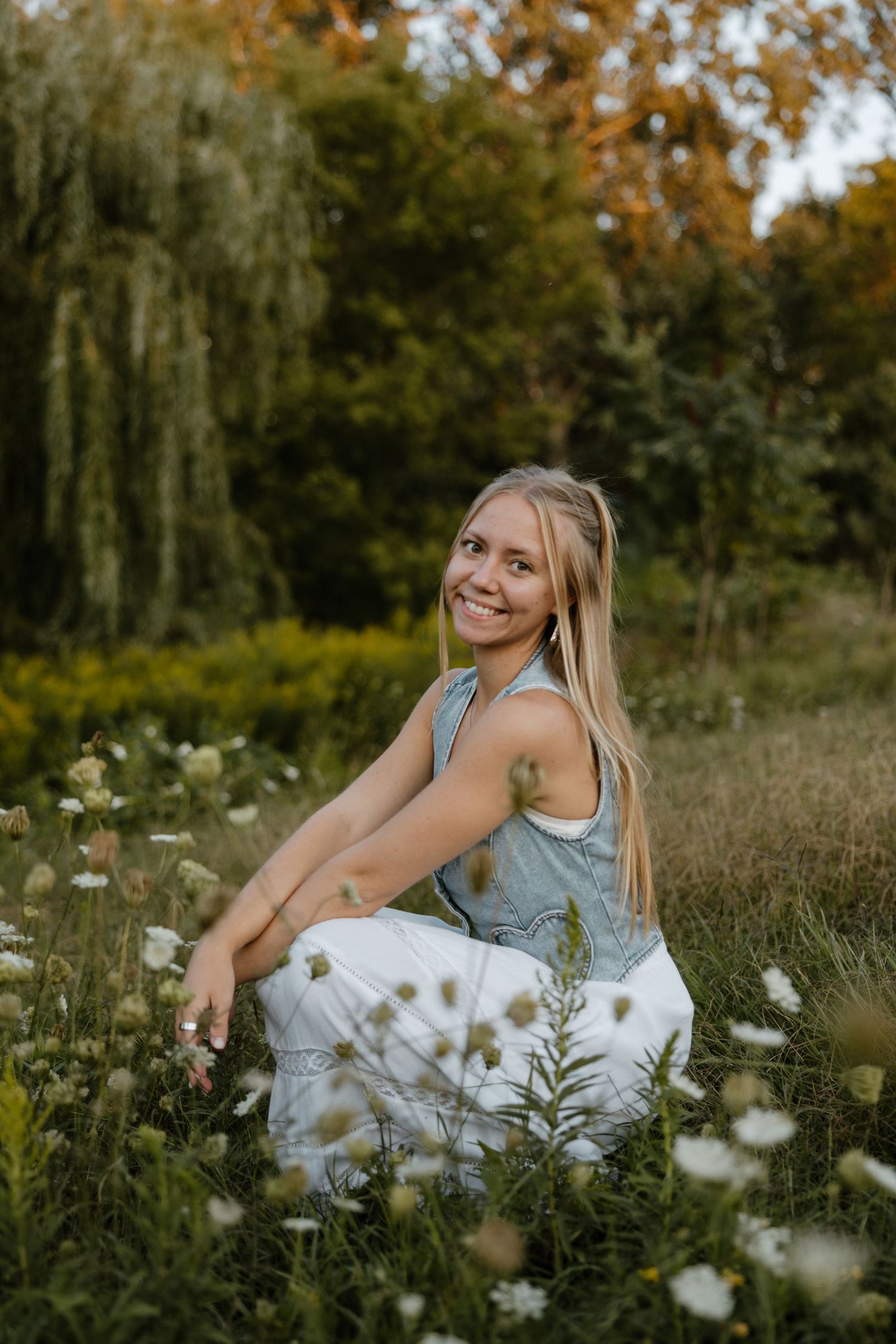 A smiling young woman sits in a field of white flowers, surrounded by lush greenery and autumn foliage. Fort Worth Texas photographer.