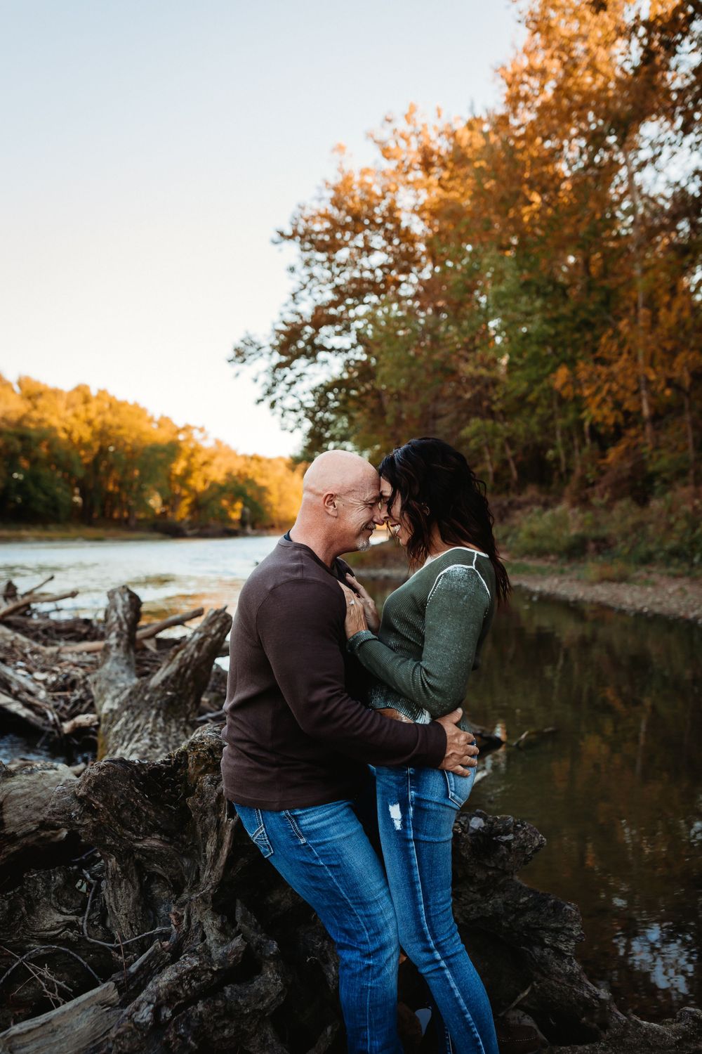Couple embraces by a tranquil lake at sunset with autumn trees reflecting golden light on the water.
