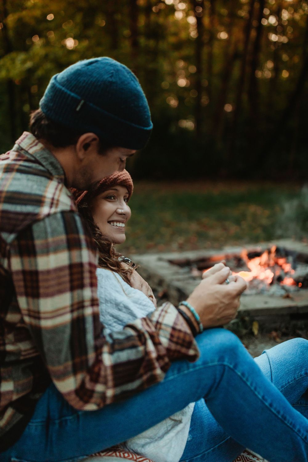 Person in plaid shirt and beanie relaxes by campfire while using phone on autumn evening.