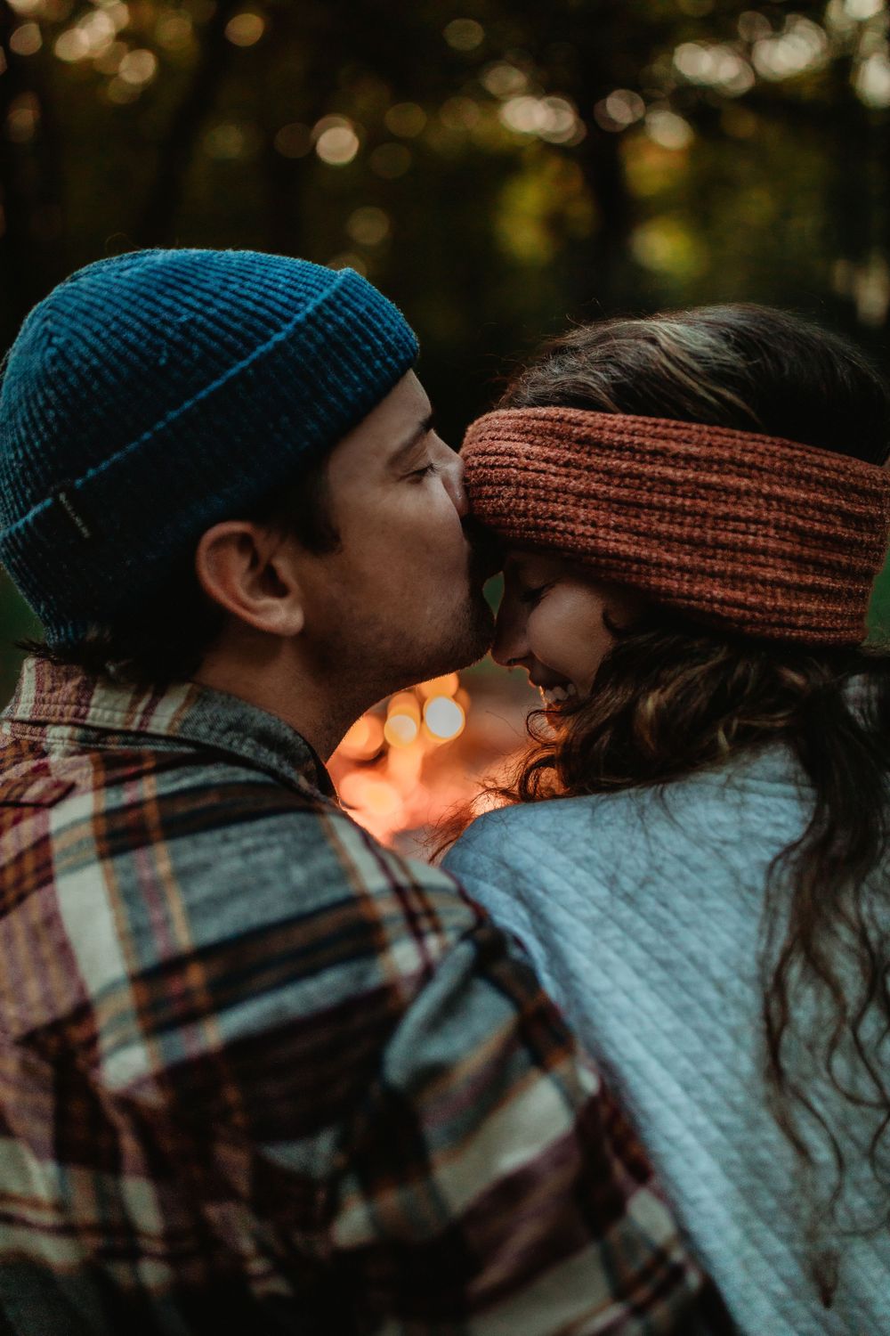 A couple wearing winter hats share an intimate moment outdoors with sunlit bokeh in the background.