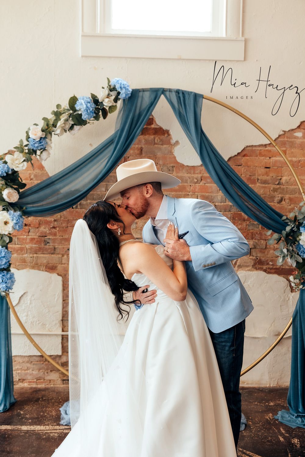 Wedding couple sharing romantic kiss under blue draped arch with floral decorations.