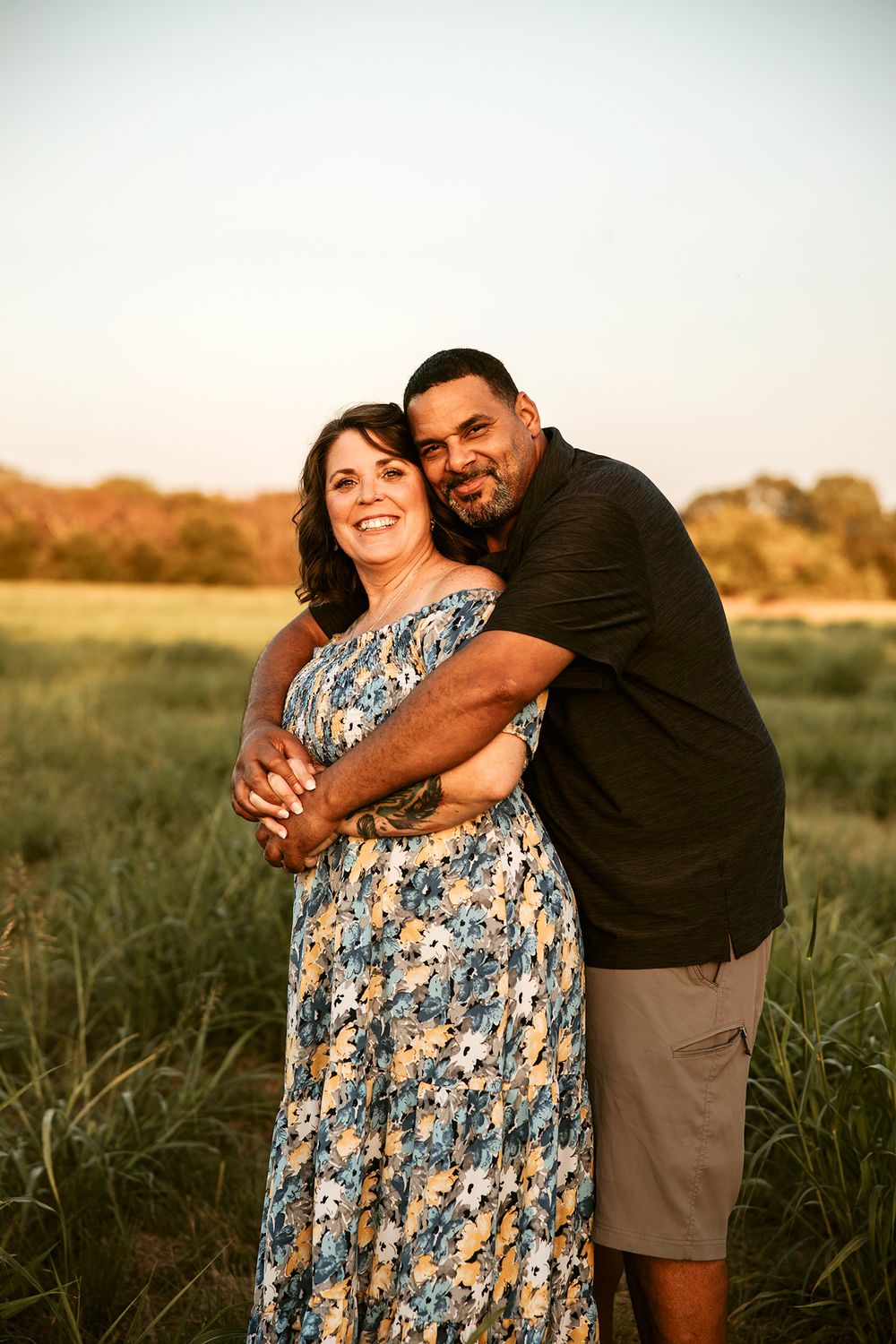 Sunset photoshoot in field with couple embracing, wearing casual summer attire.