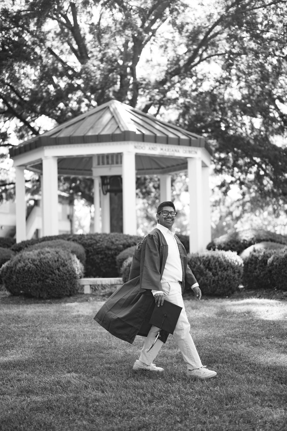 Black and white photo of a graduate in cap and gown walking on grass with a white gazebo in the background.