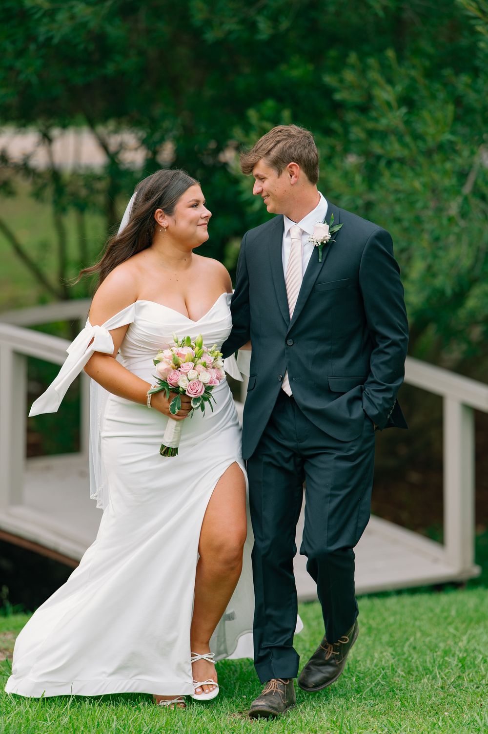 Romantic wedding couple sitting on white steps in a lush garden setting with soft pink floral bouquet.