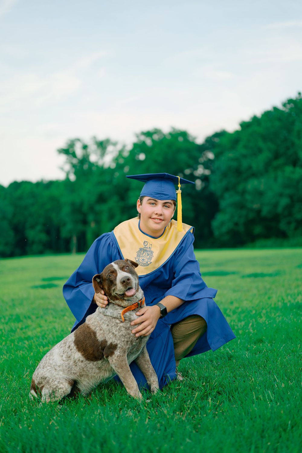 Graduate in blue cap and gown plays with brown and white dog on green grass field during celebration photoshoot.