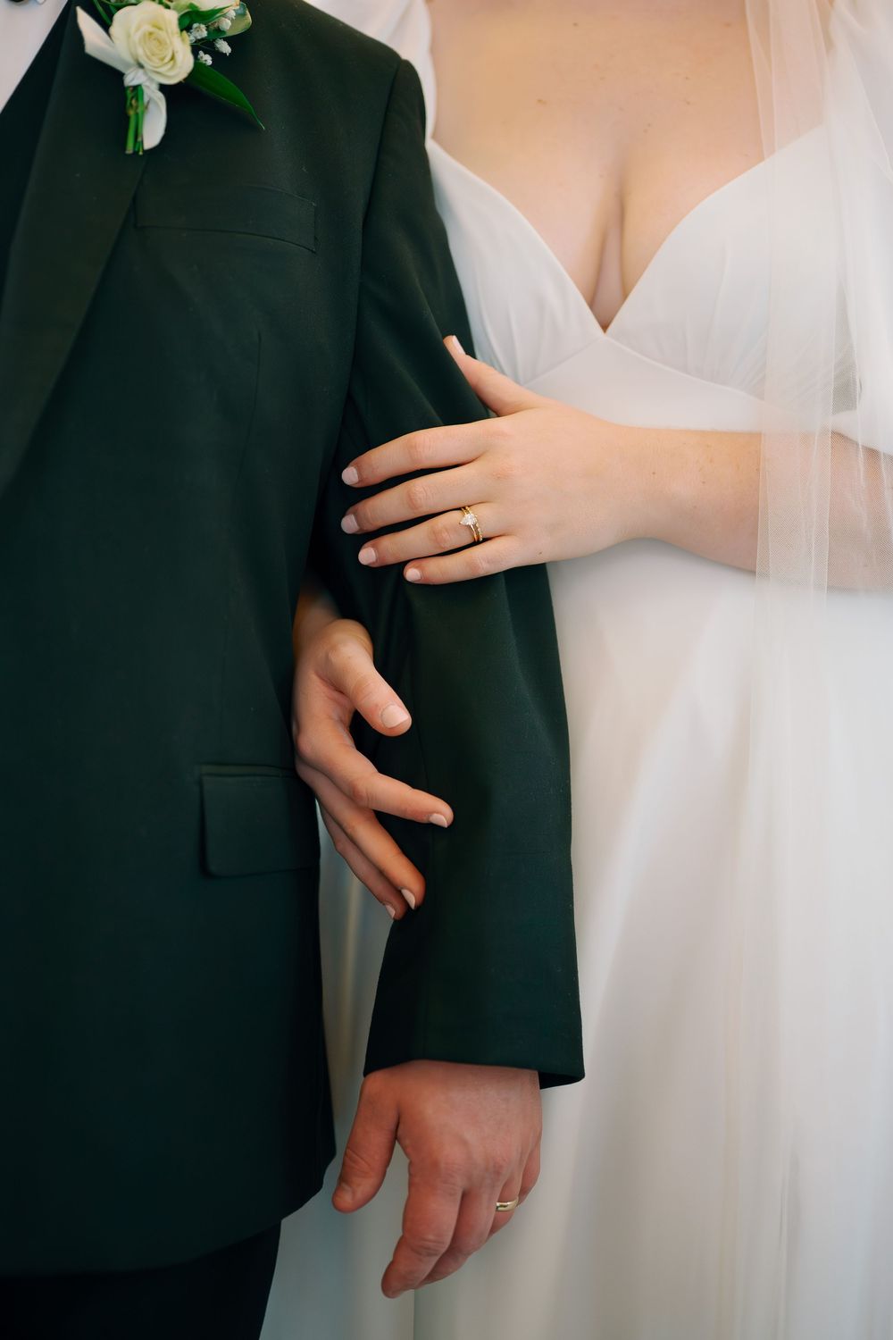 Bride in white dress with wedding ring holds groom's arm in dark green suit with white boutonniere.
