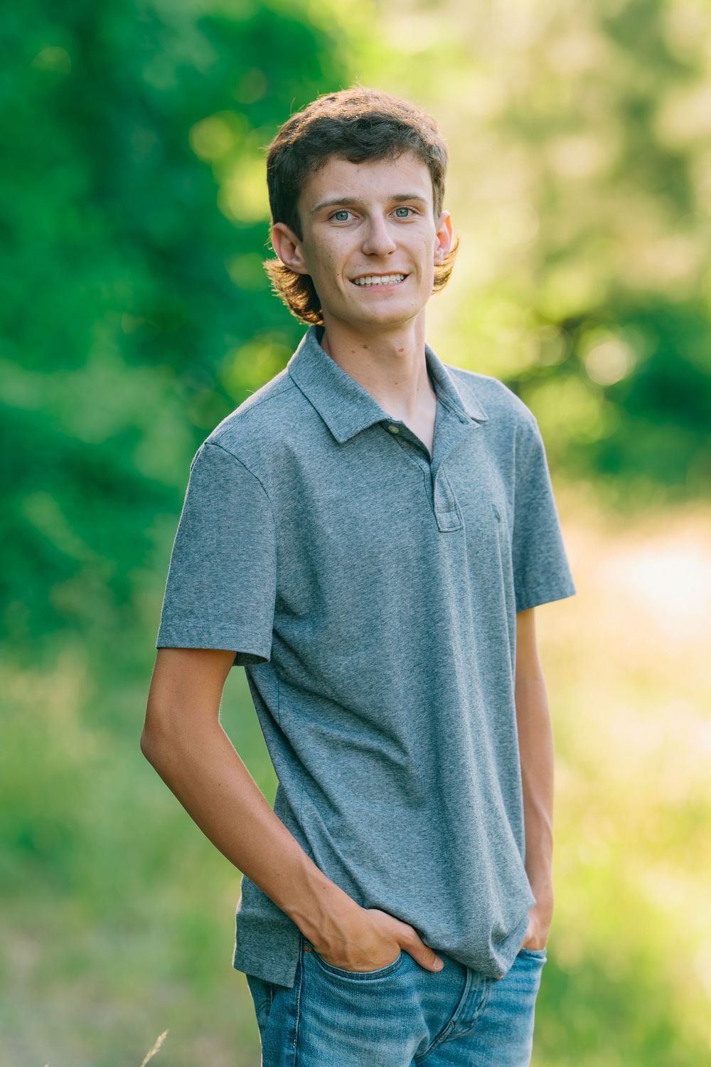 Smiling young man in gray polo shirt and jeans posing outdoors with green trees in background.