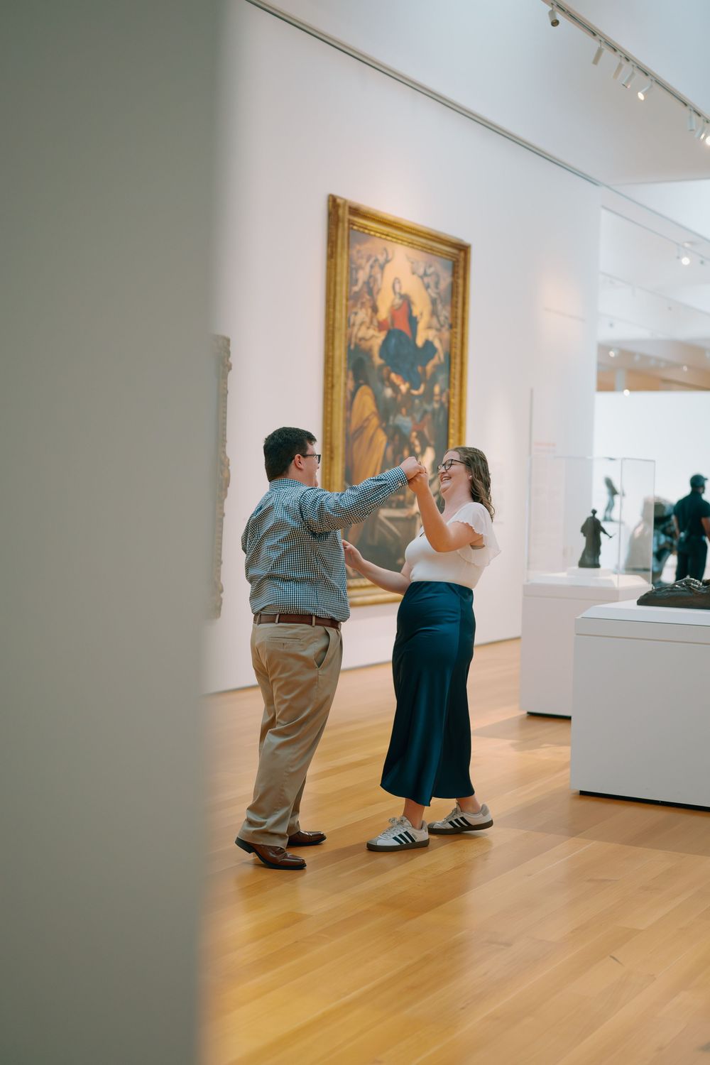A man and woman dance in an art gallery with white walls and hardwood floors, with paintings and sculptures on display.