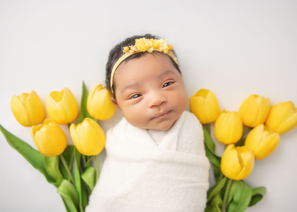 Newborn baby wrapped in white swaddle surrounded by yellow tulips wearing a floral headband.
