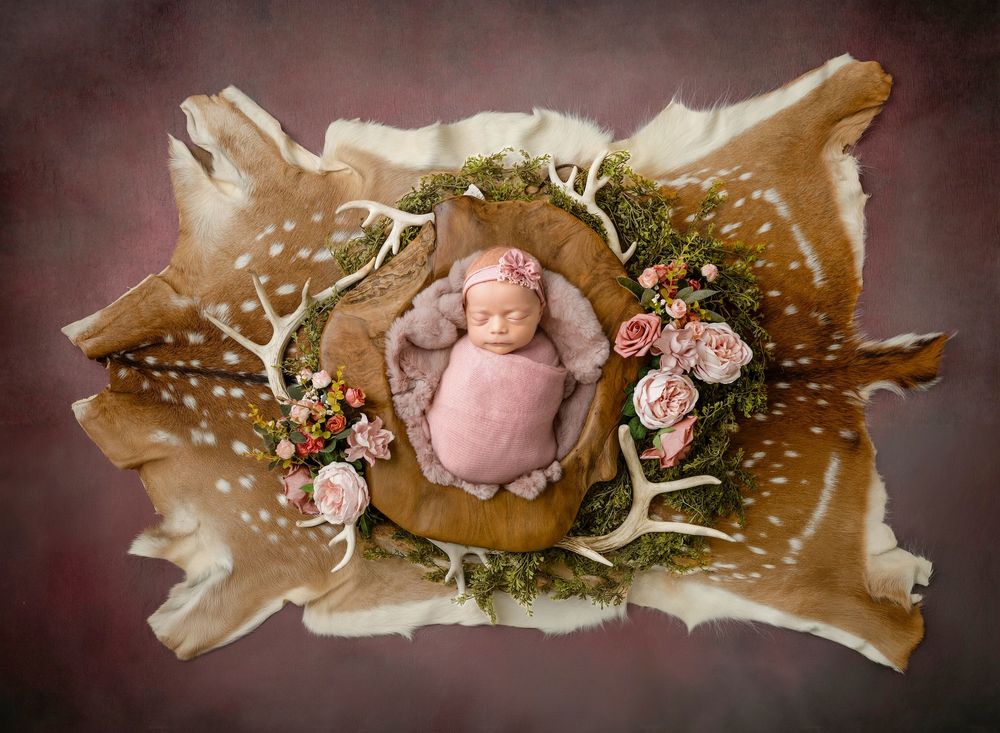 Sleeping newborn baby wrapped in pink on deer hide with floral wreath and antlers.