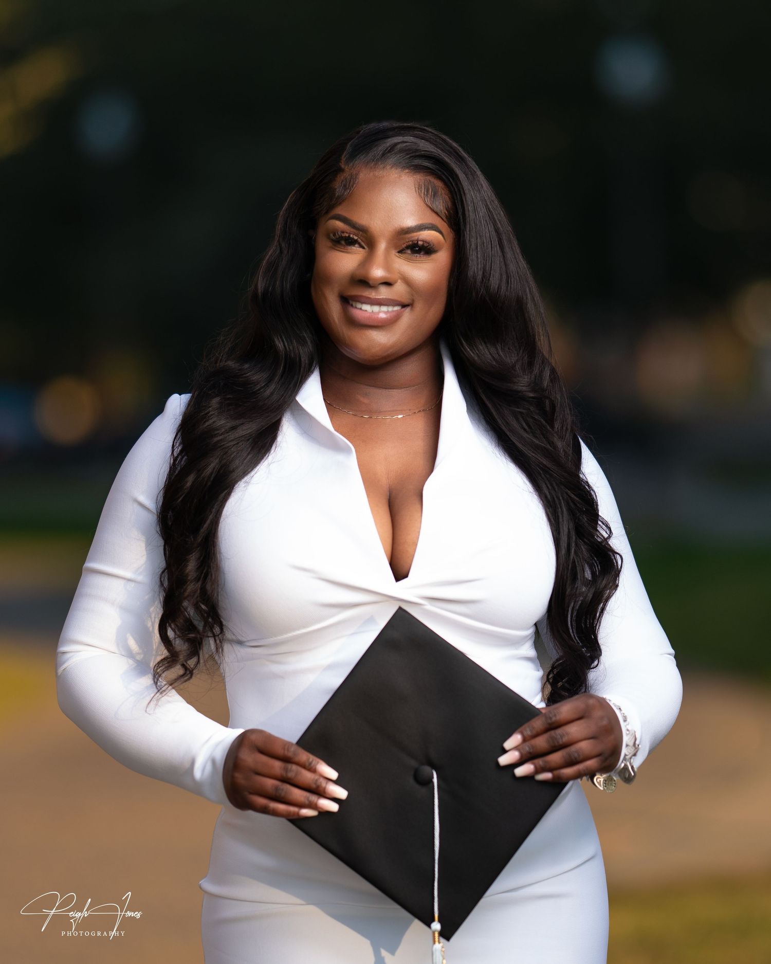 Smiling graduate in white outfit holds black graduation cap outdoors in warm golden light.