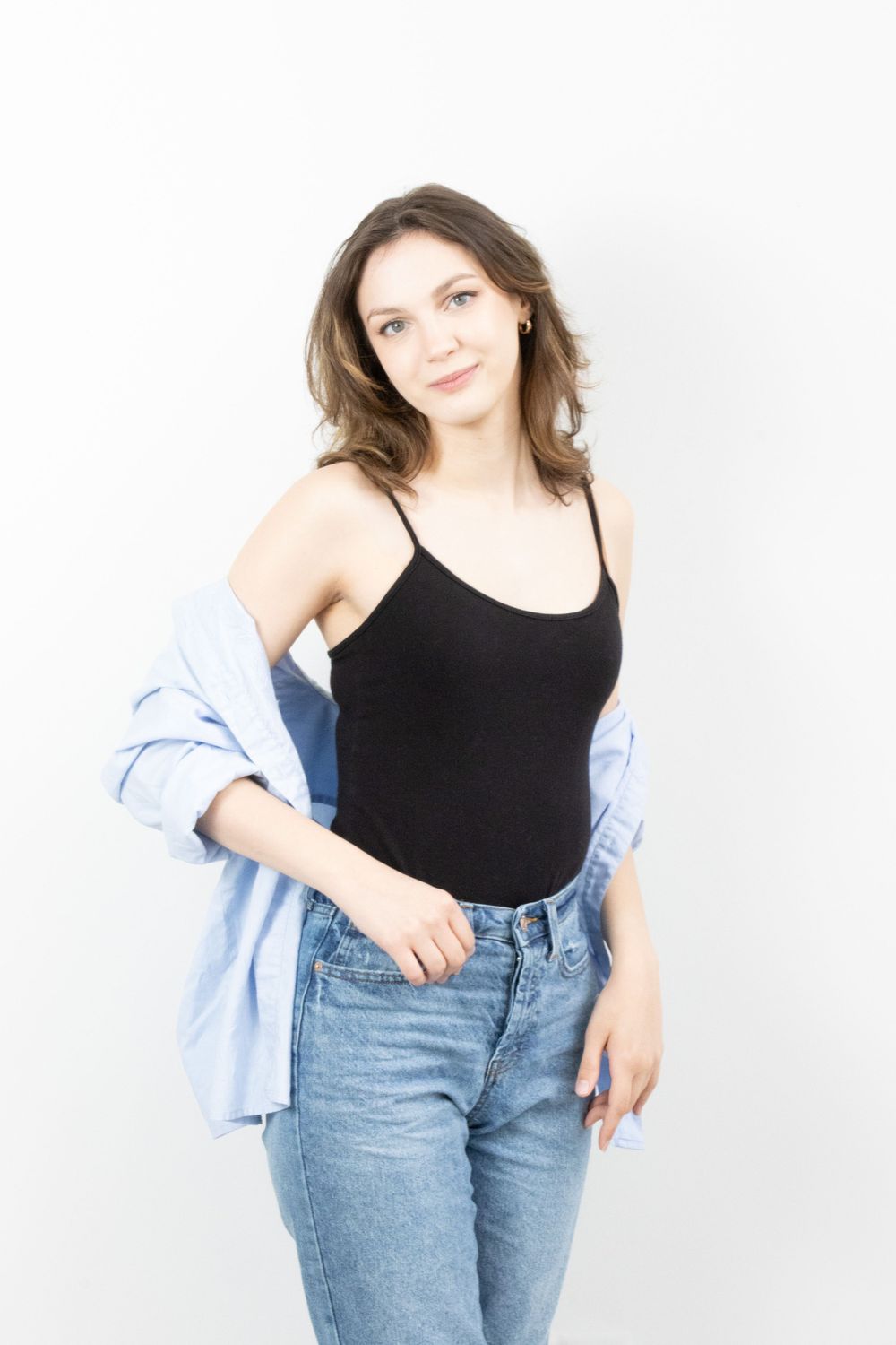 A smiling young woman in a black cami and blue shirt poses casually against a white background.