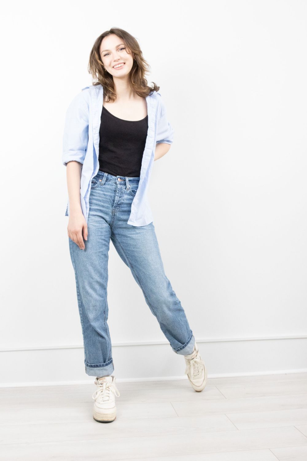 Cheerful young woman in blue shirt, black top, and jeans standing against white wall.