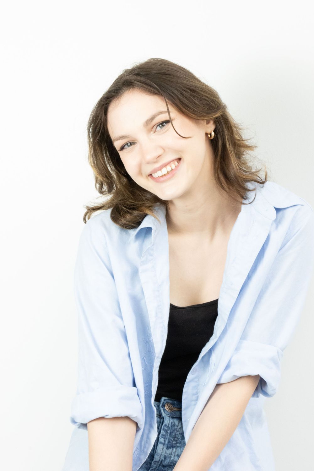 Smiling young woman with brown hair wearing blue shirt and black top on white background.