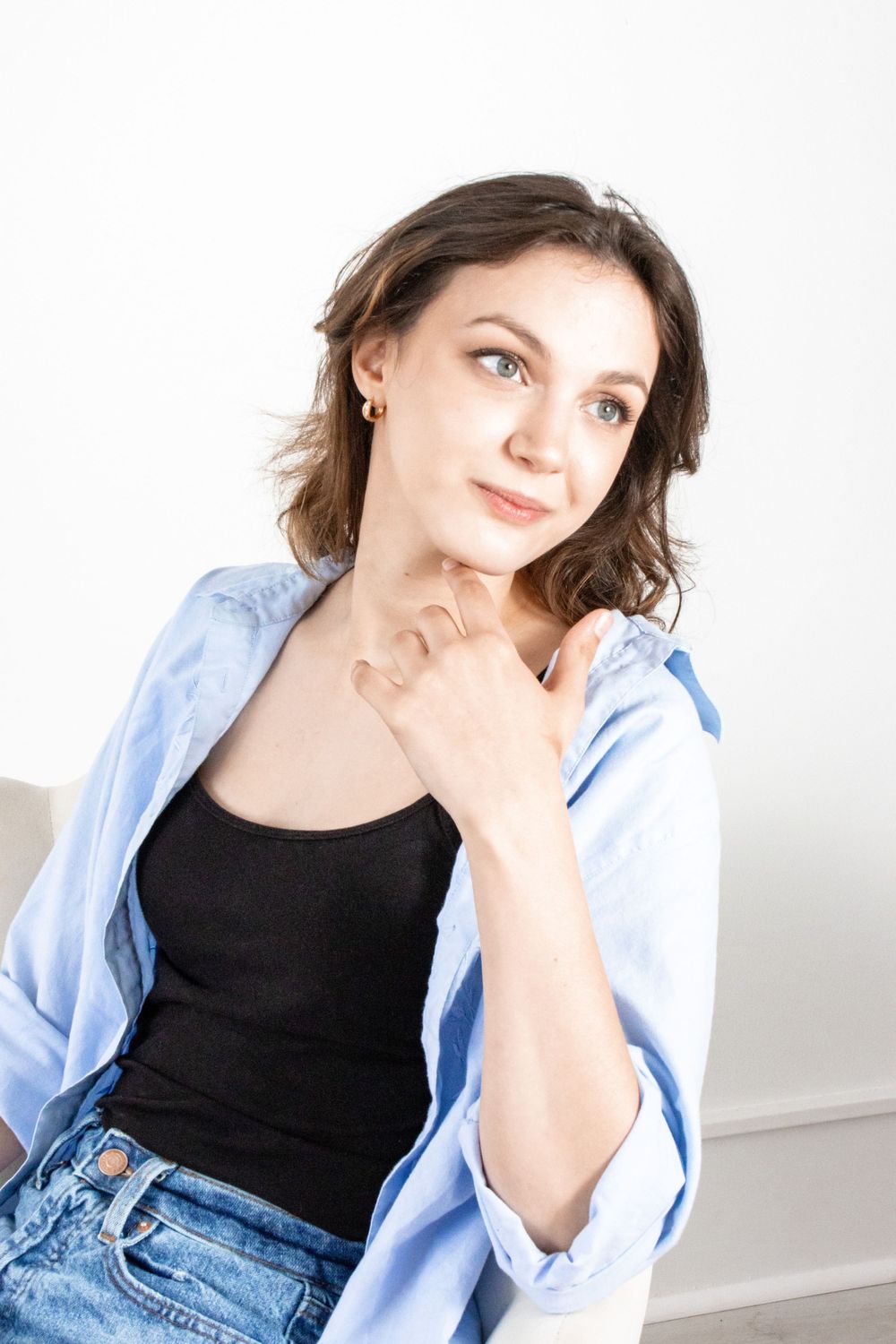 Young woman in blue shirt sitting thoughtfully with hand near chin against white background.