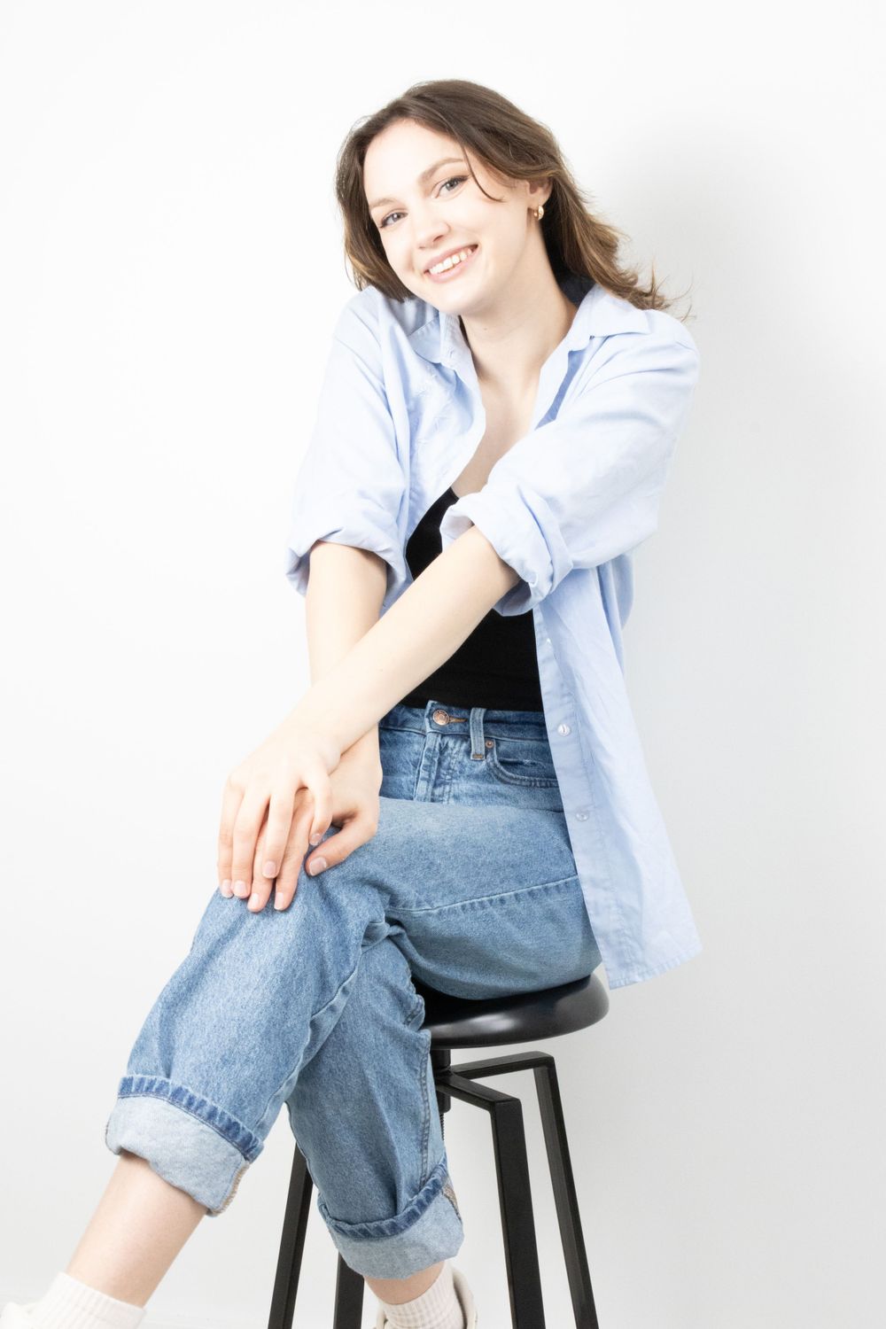 Smiling young woman sitting on black stool in blue shirt and jeans against white background.